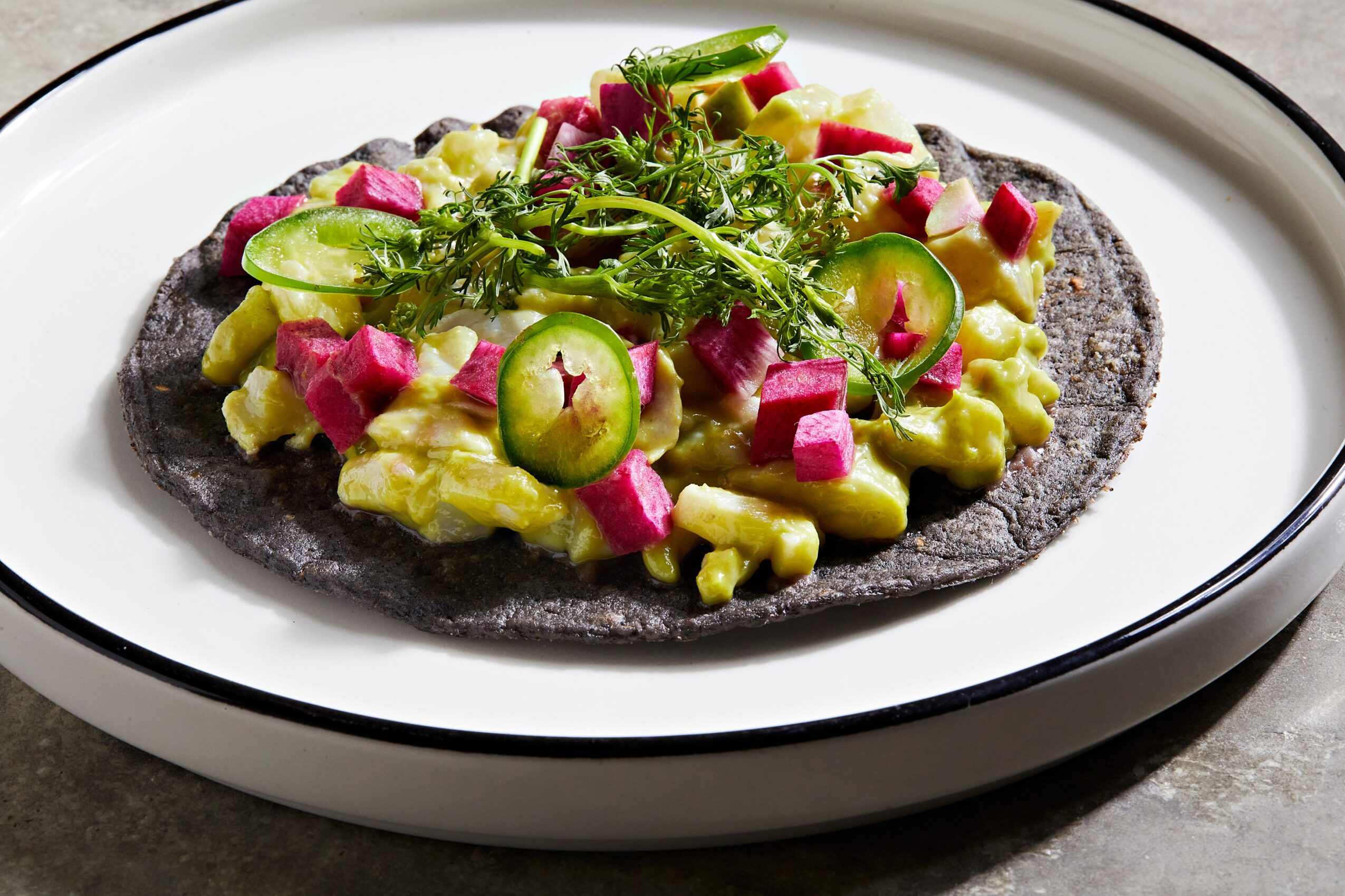 Black tortilla topped with diced cucumber, radish, and chopped herbs, garnished with fresh greens on a white plate.