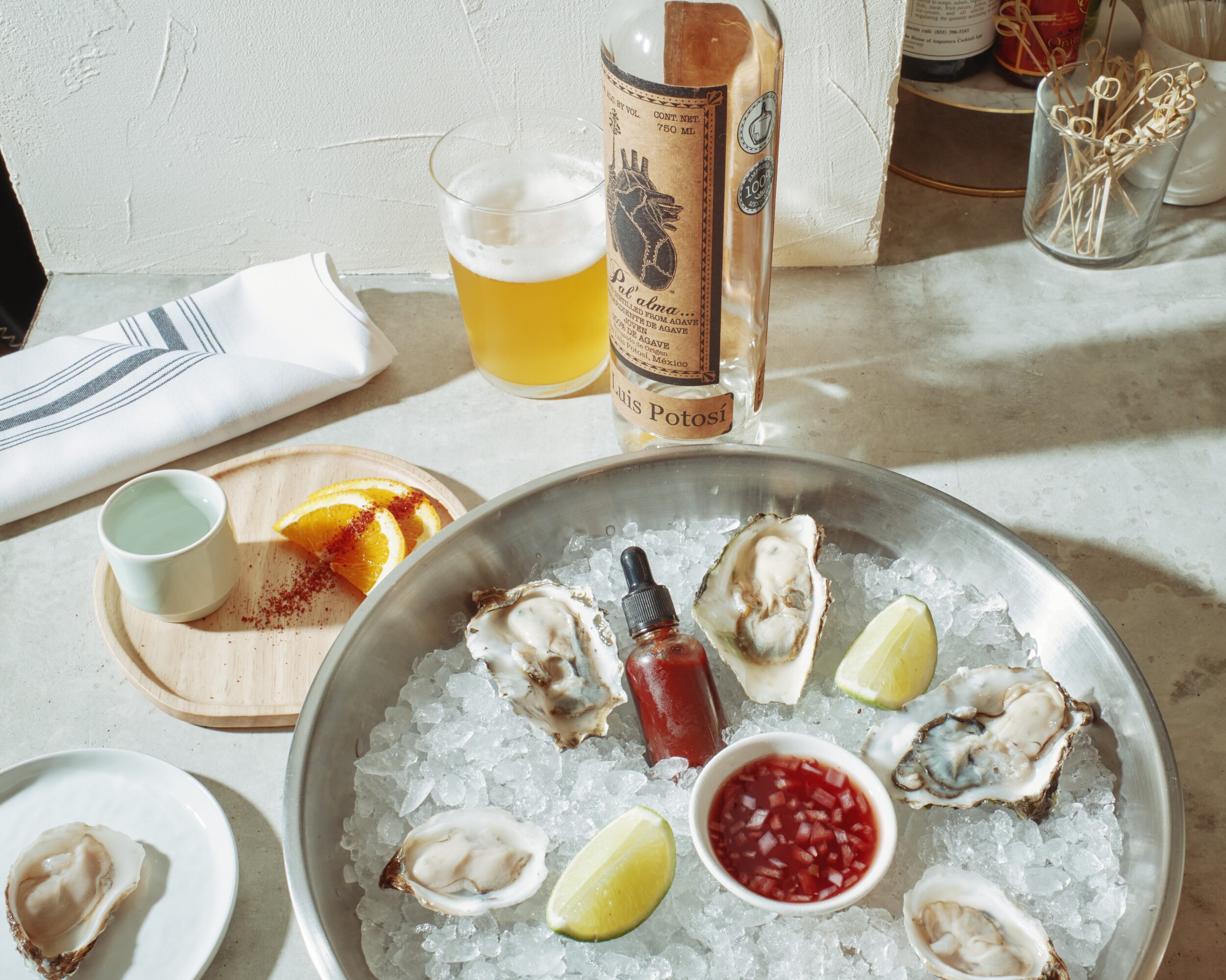 Oysters on ice with lemon wedges, a small bowl of red sauce, and a bottle of hot sauce on a table, with a glass of beer nearby.