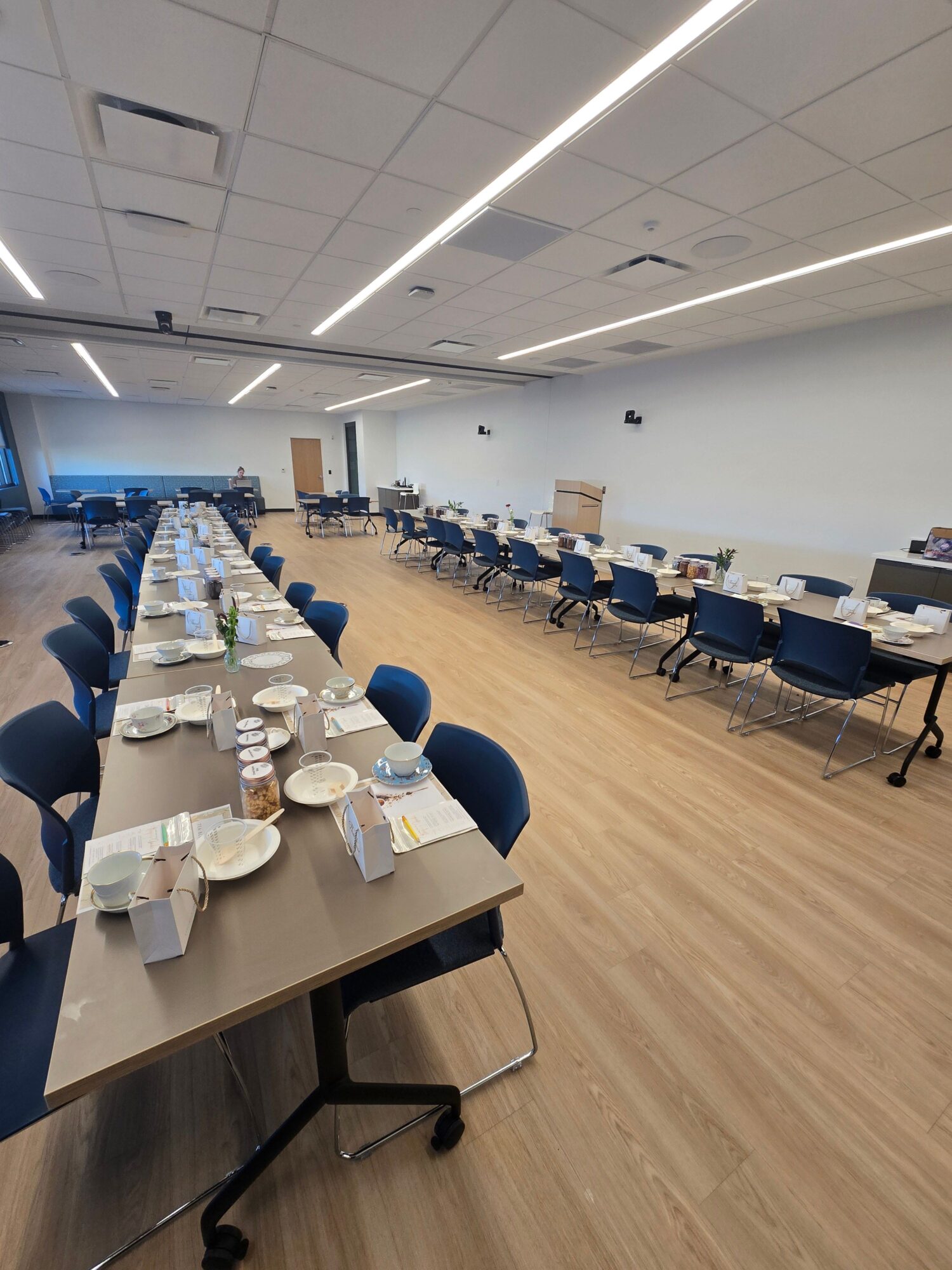 Long conference room with tables, chairs, and tableware, set up for a meeting or event, with a wooden floor and ceiling lights.