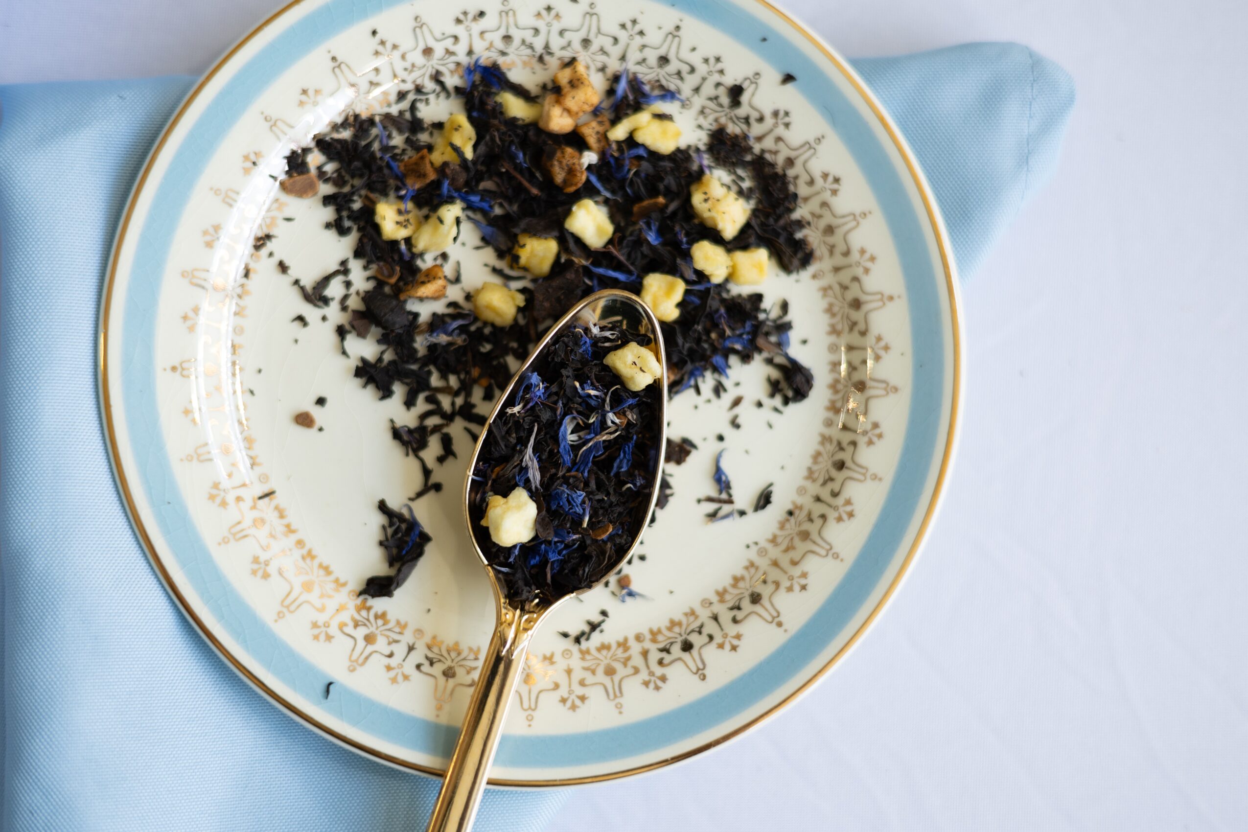A decorative plate with a gold rim holds a spoon with dried black and yellow flowers and herbs, on a light blue cloth.