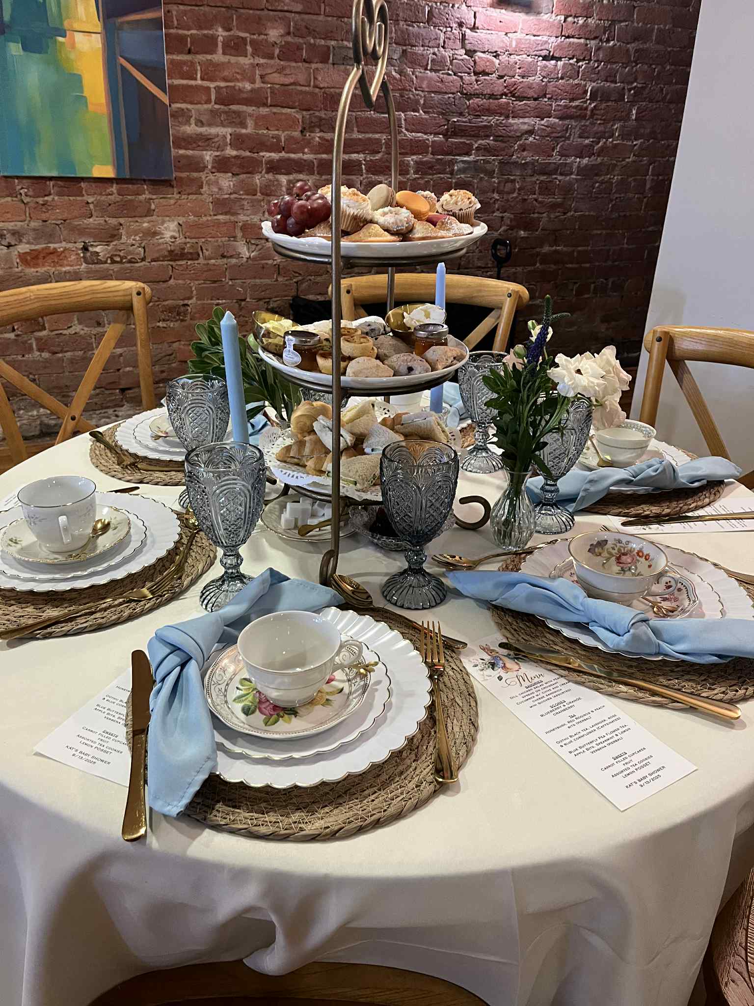 Table set with plates, cups, blue napkins, flowers, and a three-tiered tray of pastries against a brick wall.