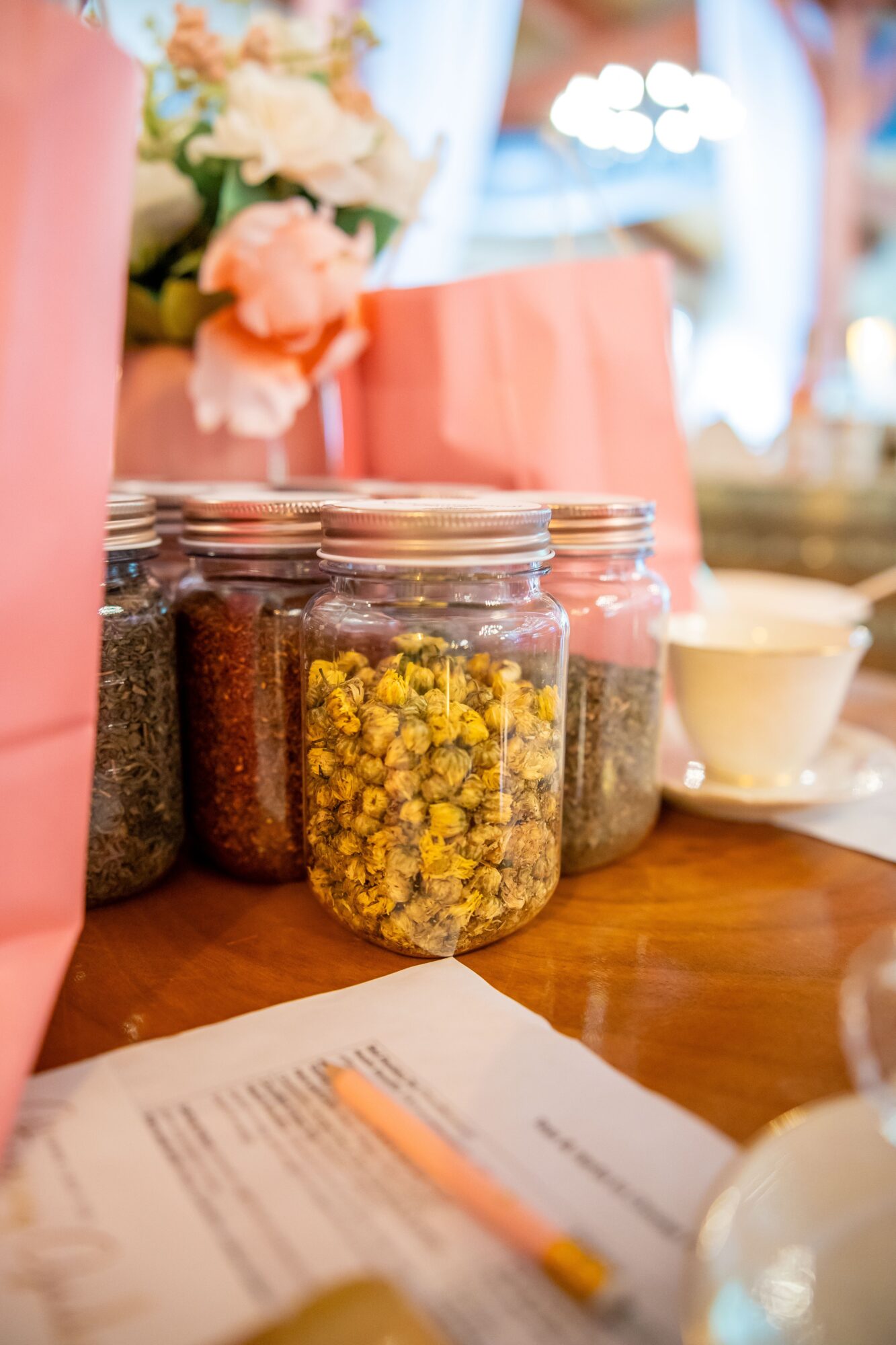 Several glass jars filled with spices or herbs on a table, with a pink bag and a flower arrangement in the background.