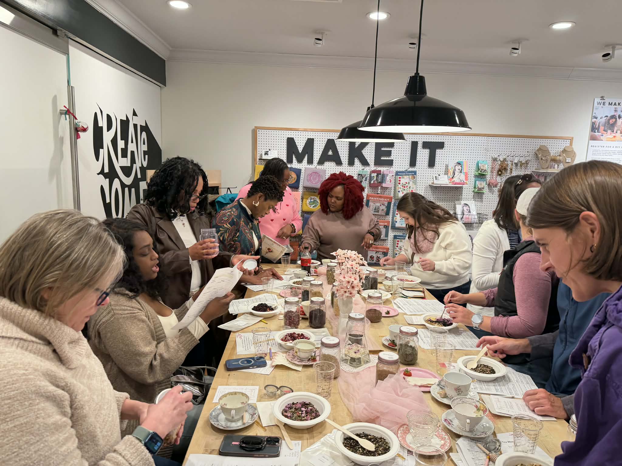 Group of women gathered around a table, working on craft projects in a room with a 'MAKE IT' sign on the wall.