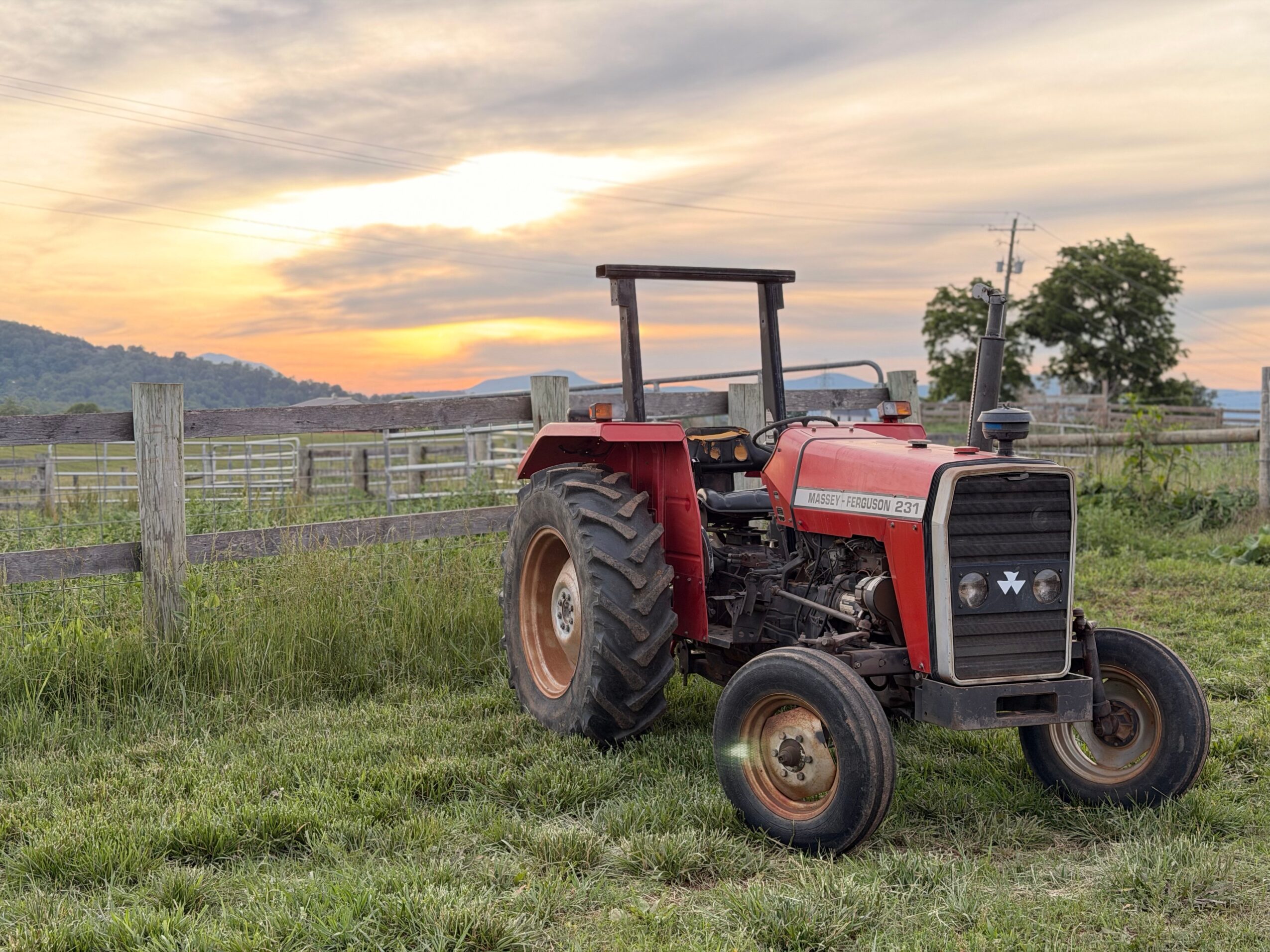 Red tractor parked on grassy field with a wooden fence and trees in the background, sunset sky overhead.