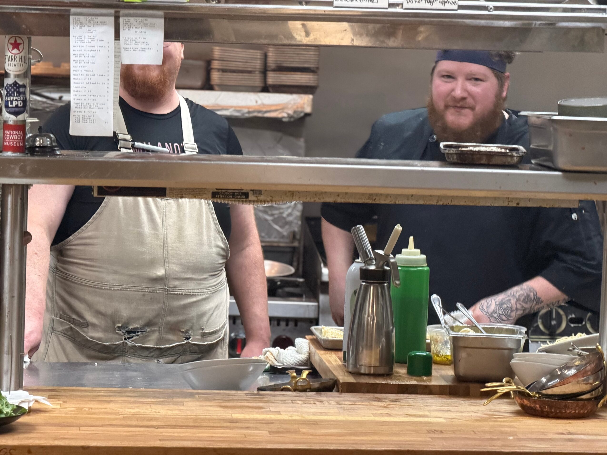 Two people behind a kitchen counter, one wearing an apron and the other with a beard, in a restaurant setting.