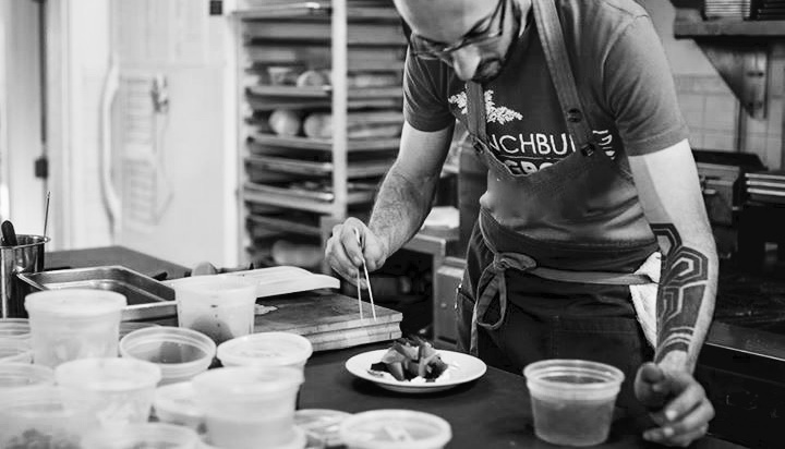 Person with glasses and tattoos preparing food in a kitchen, surrounded by bowls and utensils.