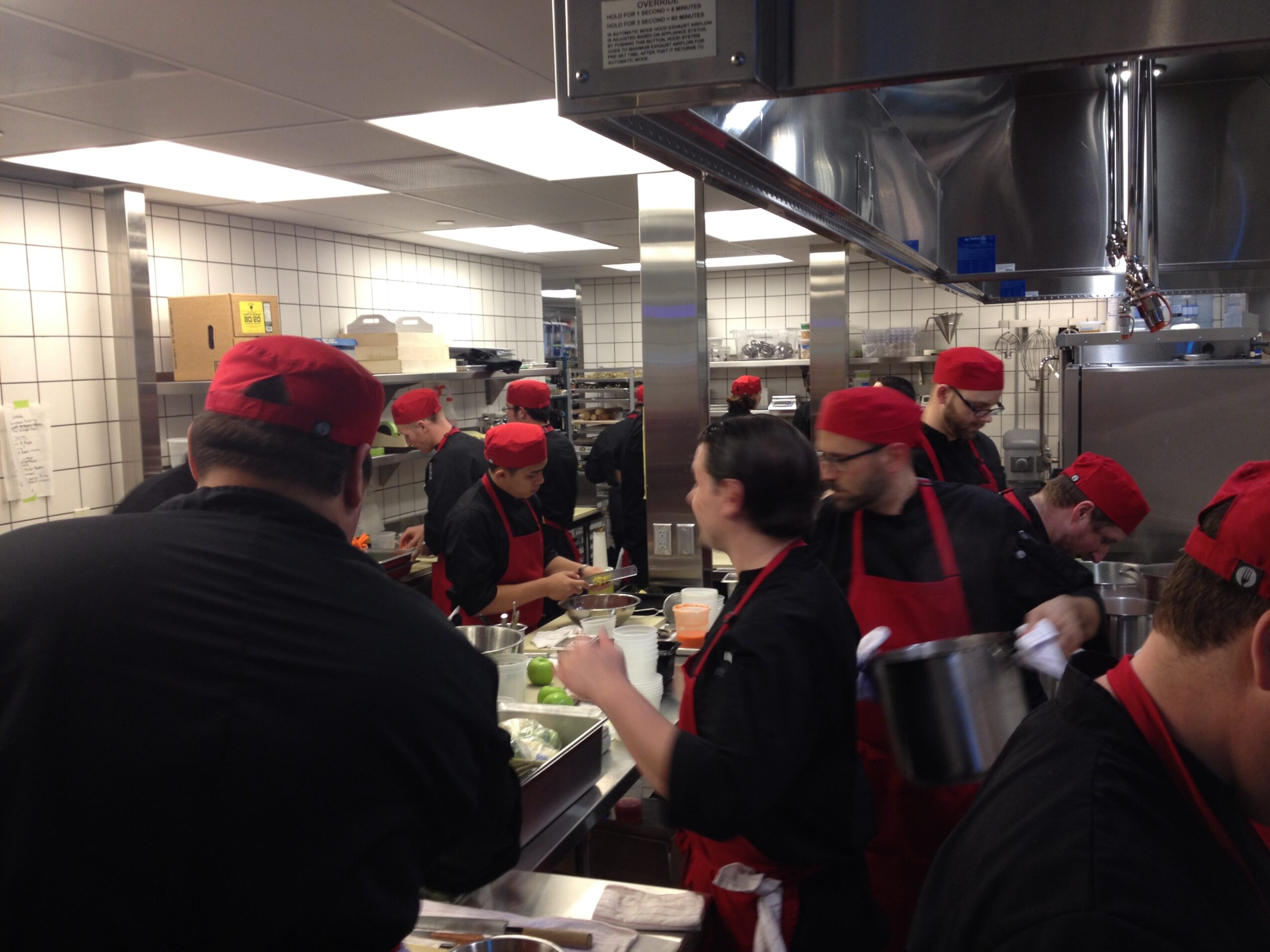 Kitchen staff wearing red hats and black uniforms work together in a busy kitchen environment.