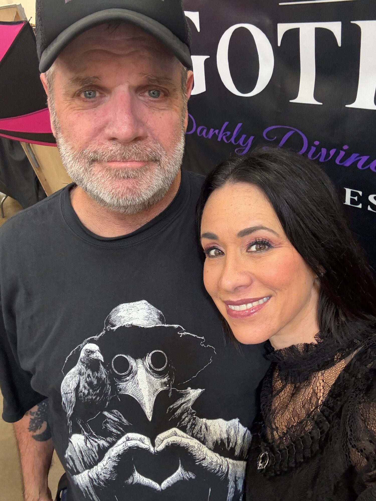 A woman with dark hair and makeup, smiling, leaning on a man with a beard, wearing a black T-shirt, in front of a sign.