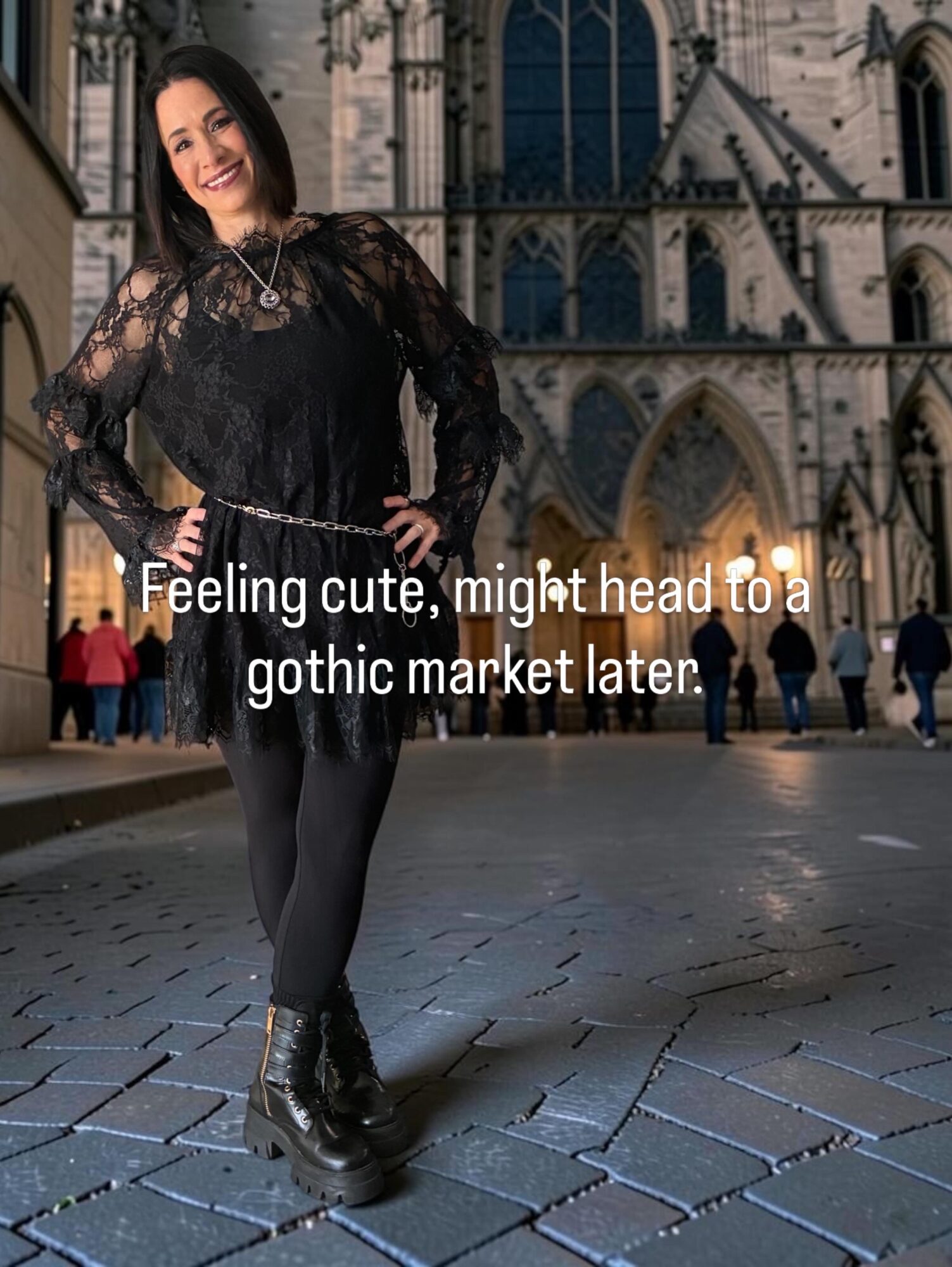 Young woman in gothic attire standing on cobblestone street in front of a cathedral, smiling, with people in background.