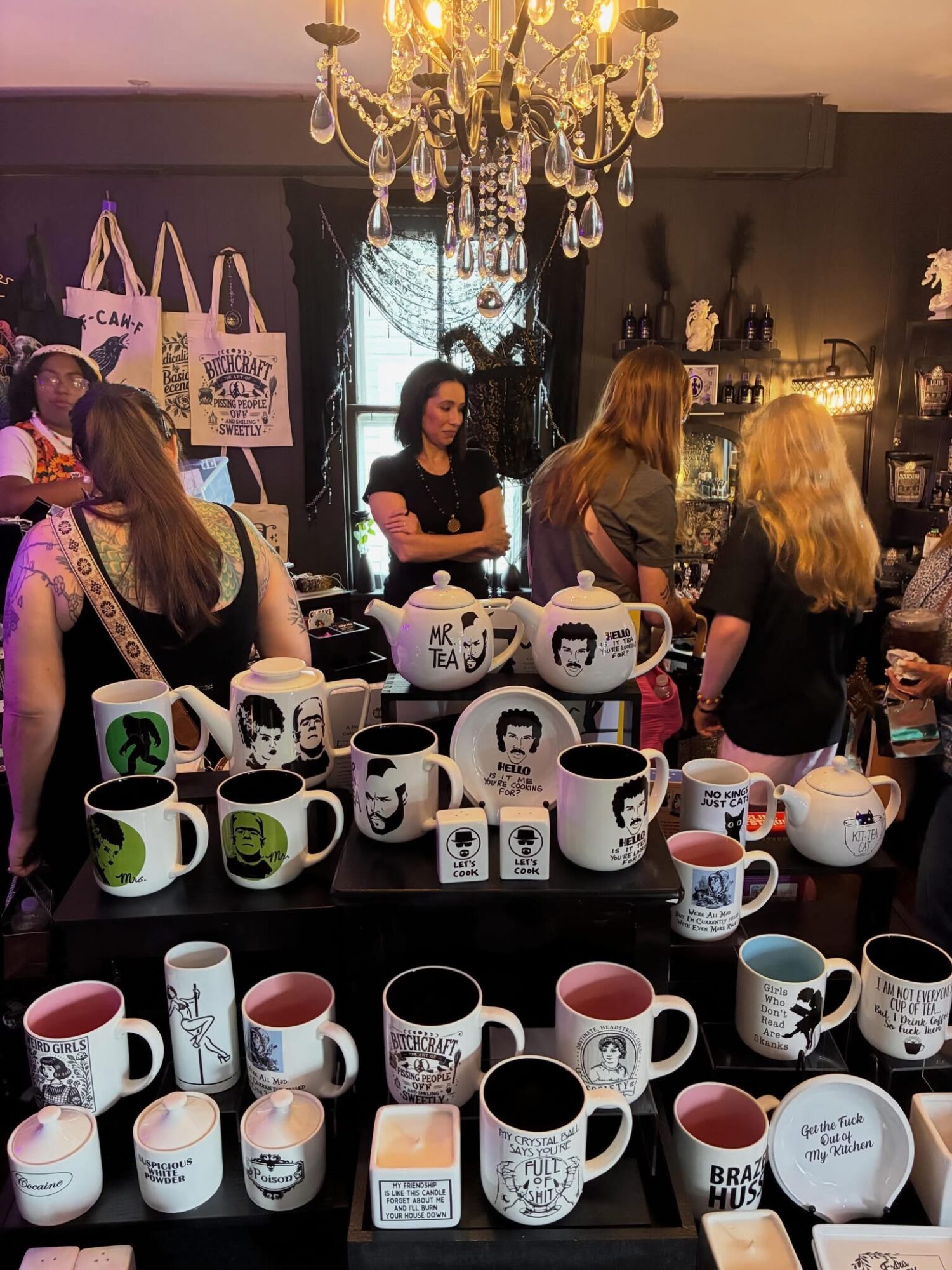 People browsing mugs and tote bags in a shop with a chandelier, shelves, and a window in the background.