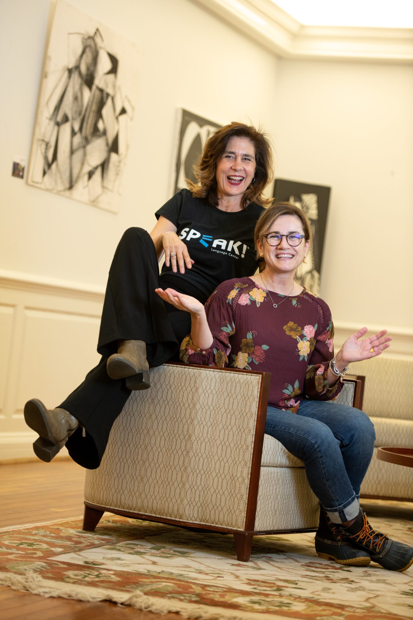 Two women smiling and sitting on a chair in an art gallery, with artwork on the wall behind them.