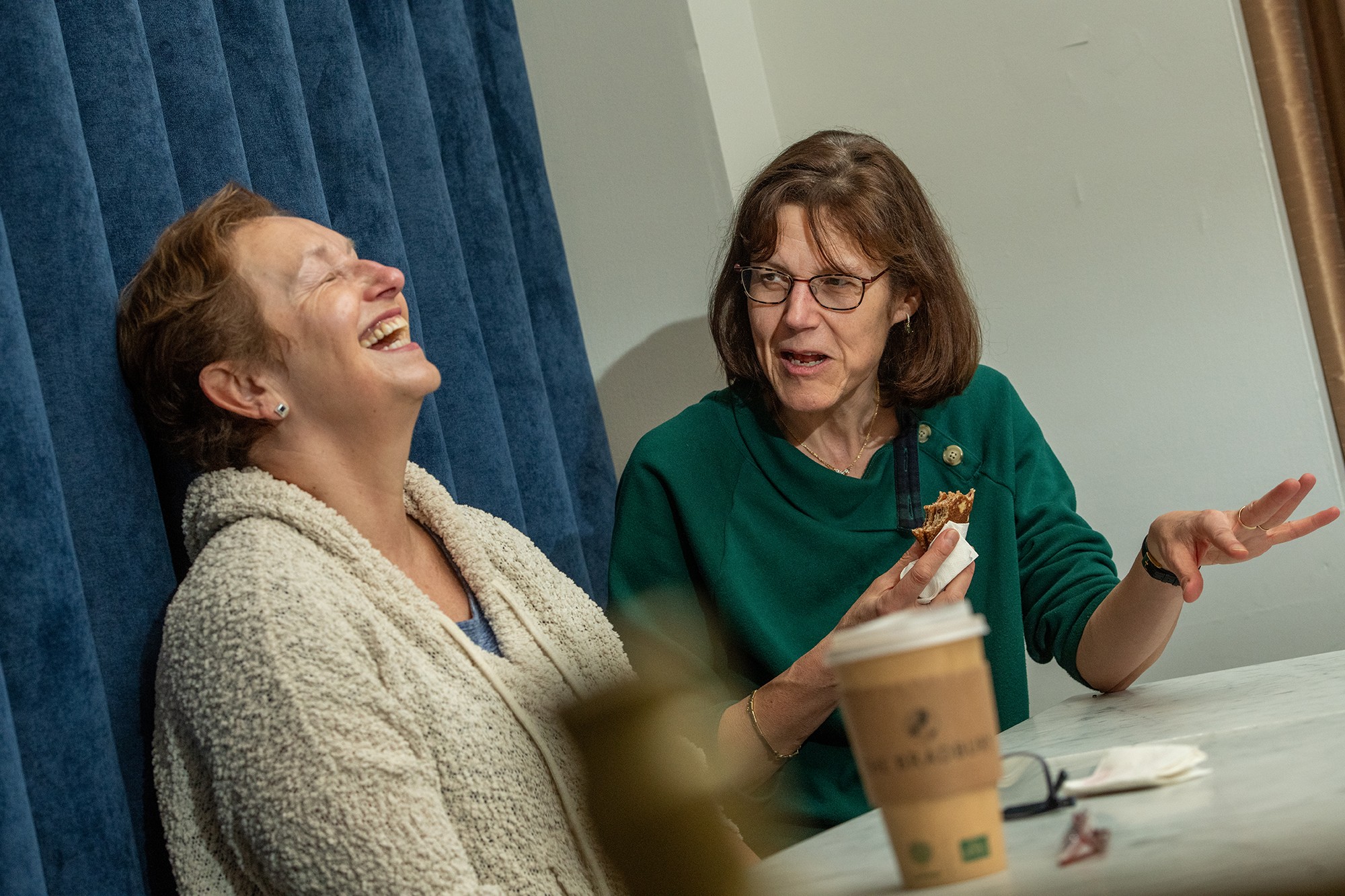 Two women sit at a table, one laughing and the other speaking, with a coffee cup in front of them.