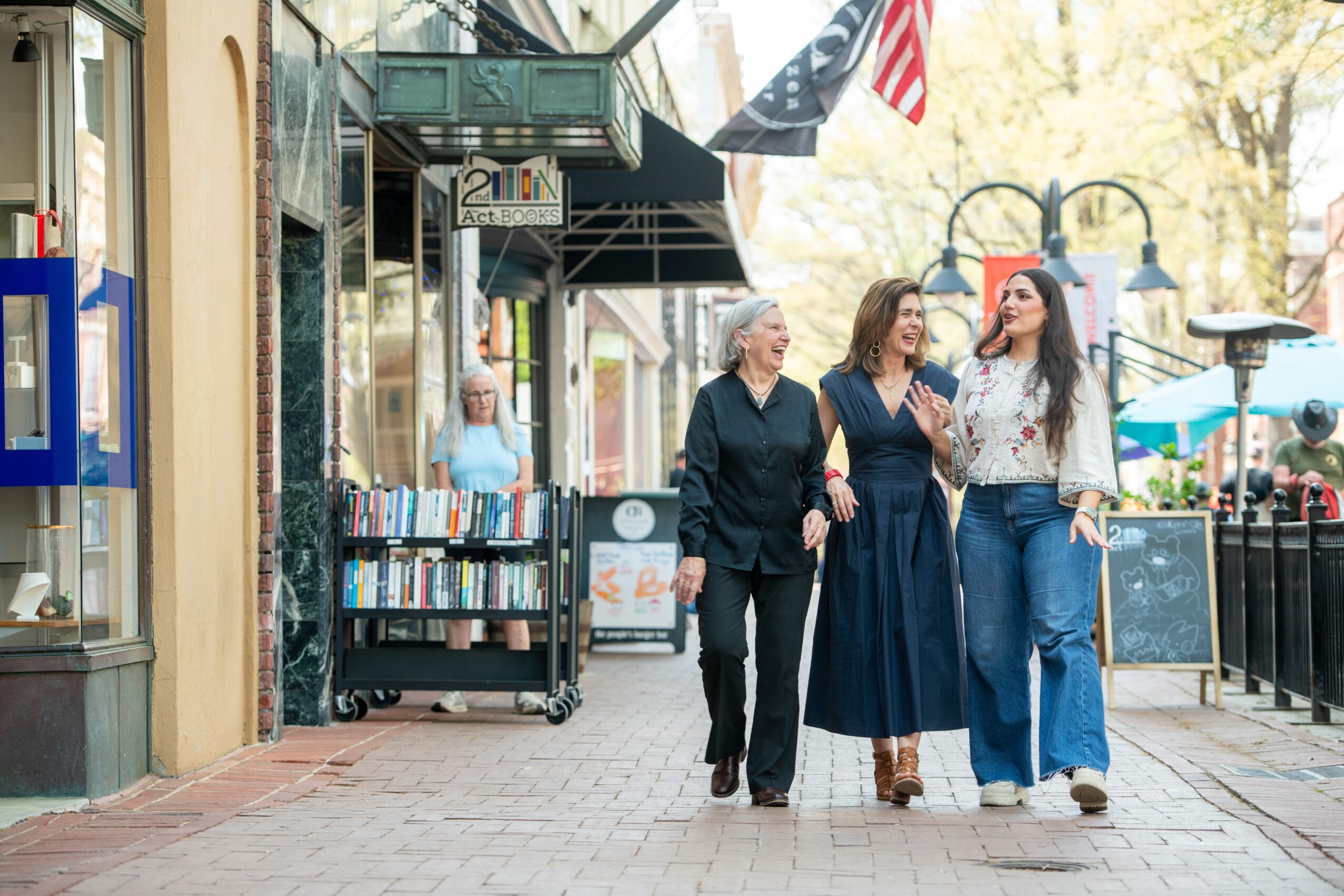 Three women walk together on a city sidewalk, talking and smiling, with shops and flags in the background.