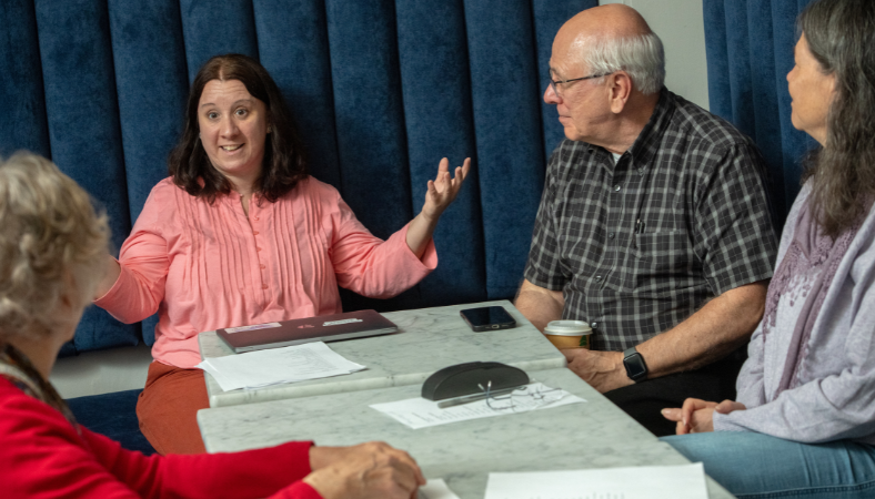 Four people sit around a table engaged in conversation, with a woman speaking animatedly, papers and a phone on the table.