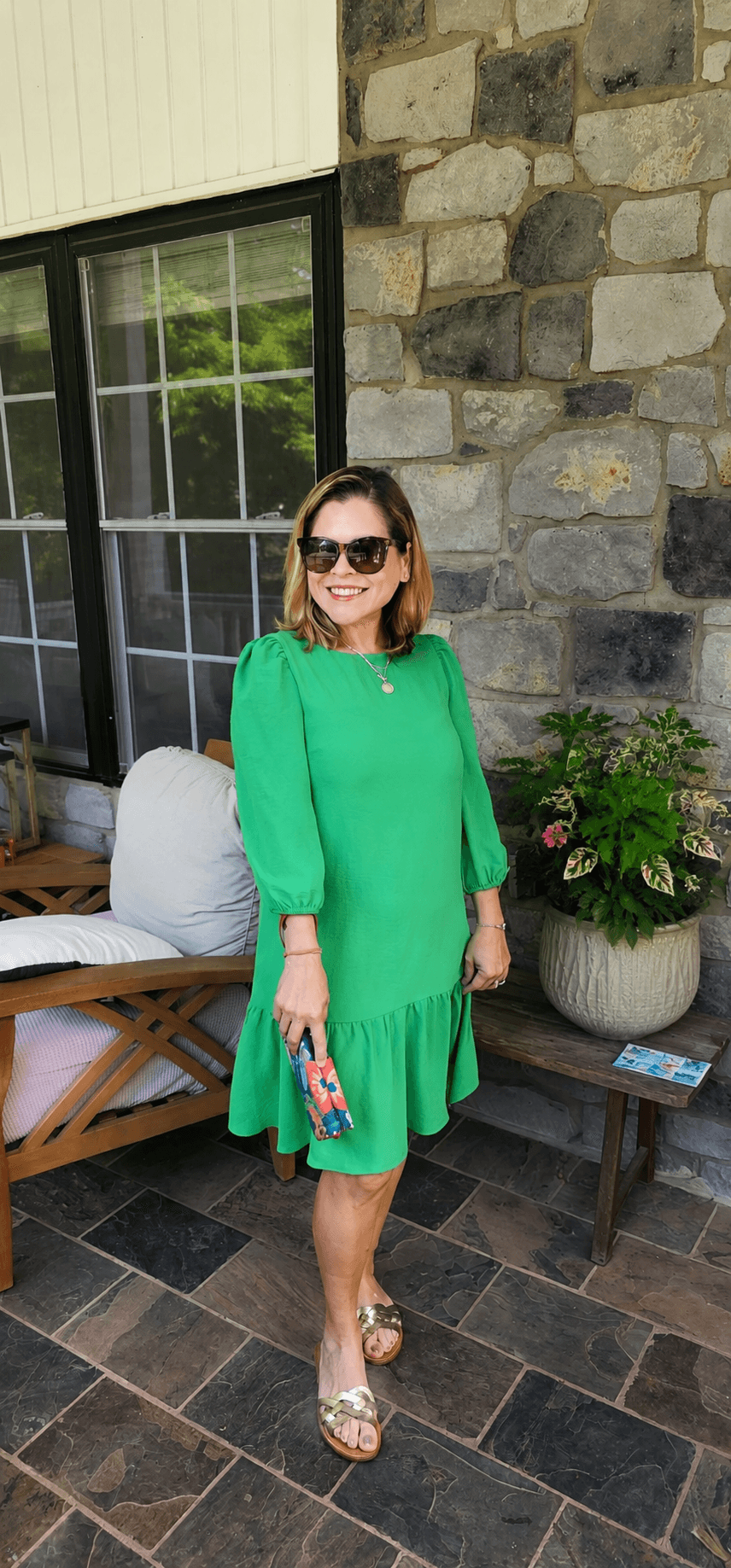 Woman in green dress and sunglasses standing outside near a stone wall and window, holding a small bag, smiling.