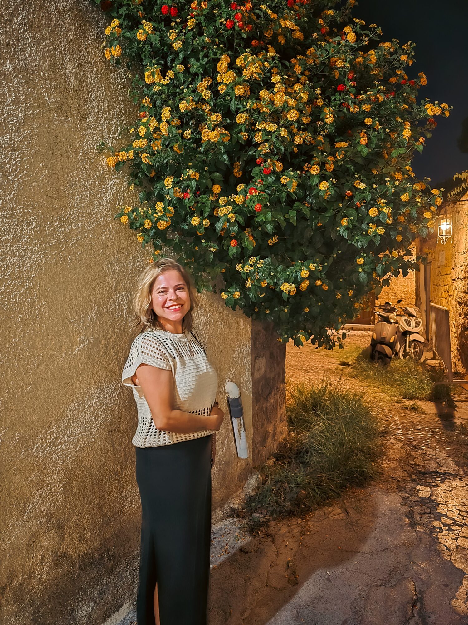 Woman smiling standing next to a large flowering bush against a wall at night.