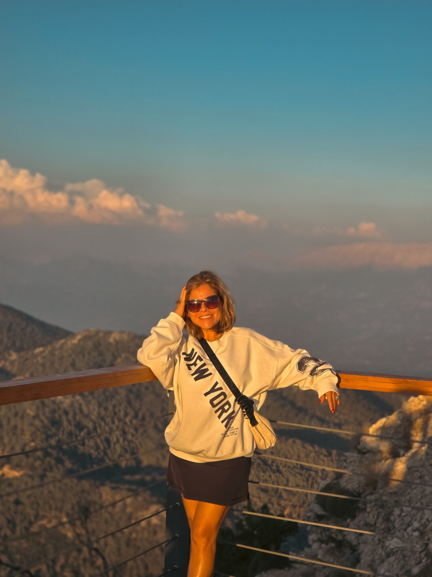 Woman in sunglasses and a white sweatshirt with black text, standing outdoors on a mountain with a railing, sunset sky in background.