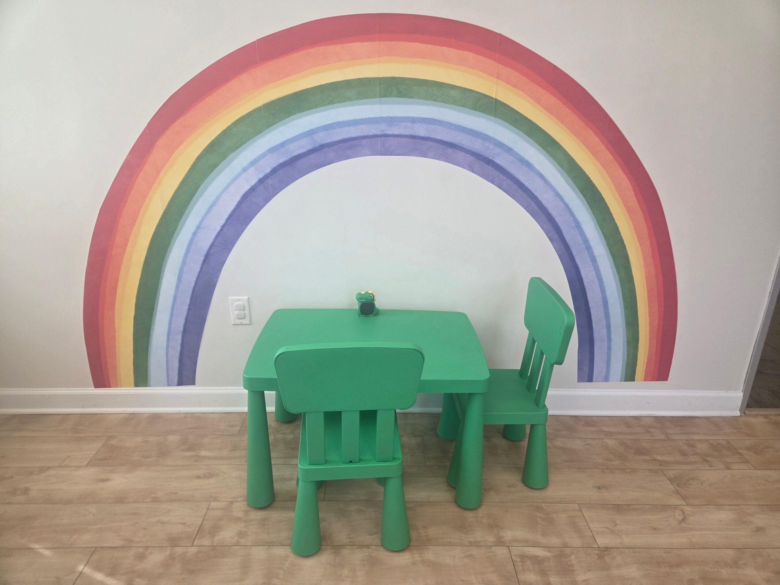 Green table with two matching chairs in front of a rainbow mural on a white wall, wooden floor.