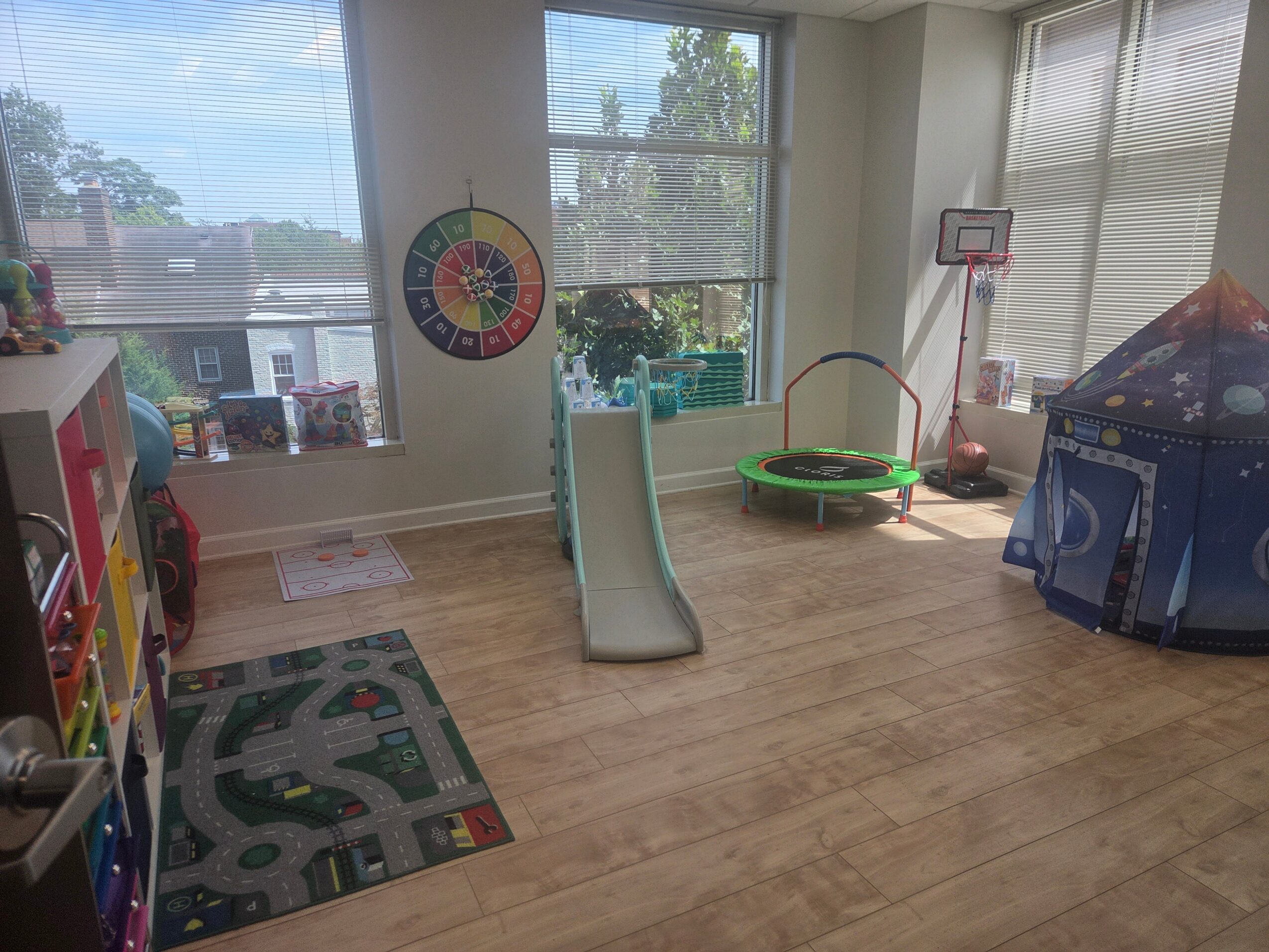 Indoor playroom with slide, basketball hoop, tent, and colorful rug, sunlight streaming through large windows.