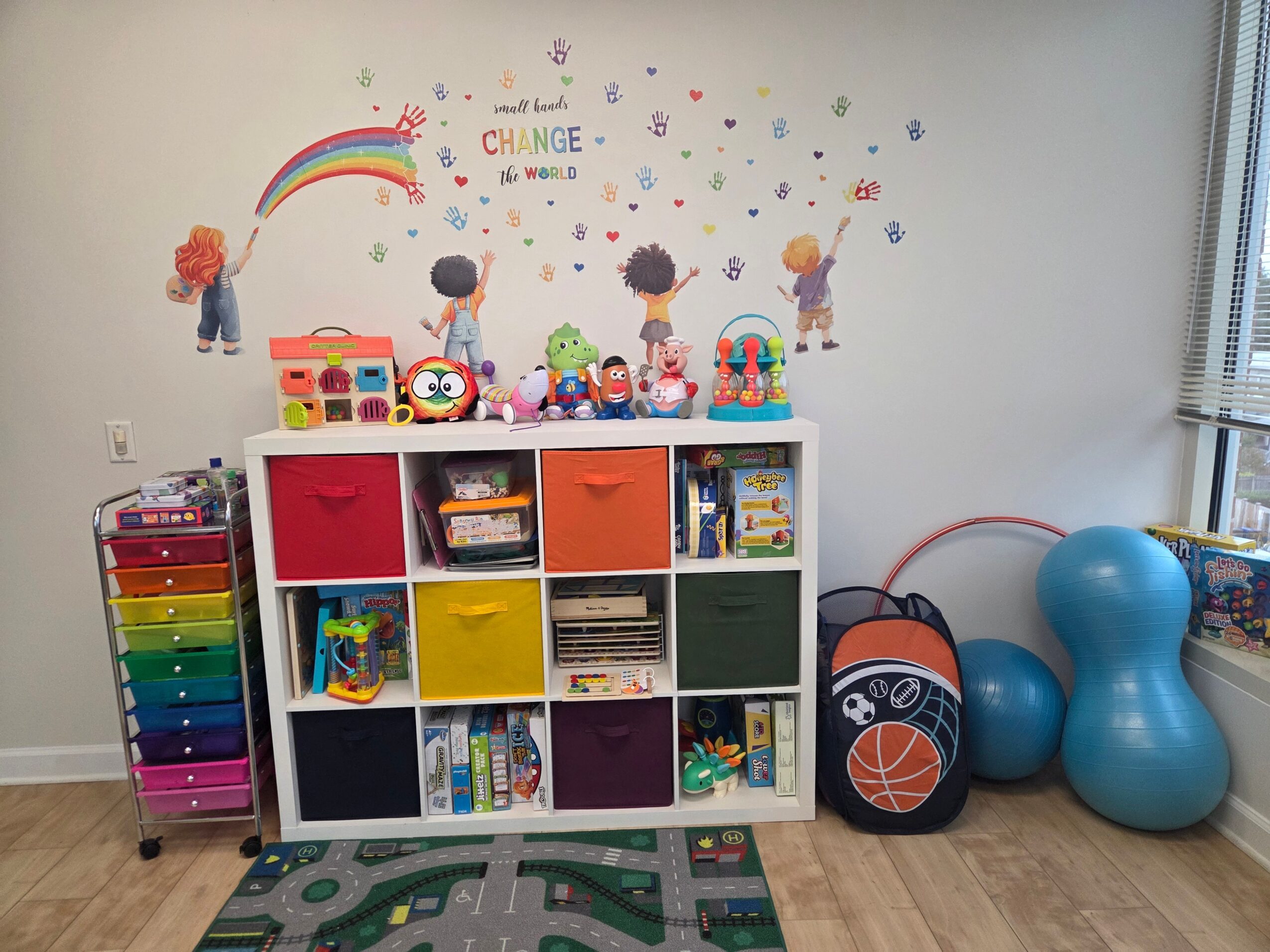 Colorful storage unit with toys and books, wall with rainbow and children, sports equipment, and large blue exercise balls.