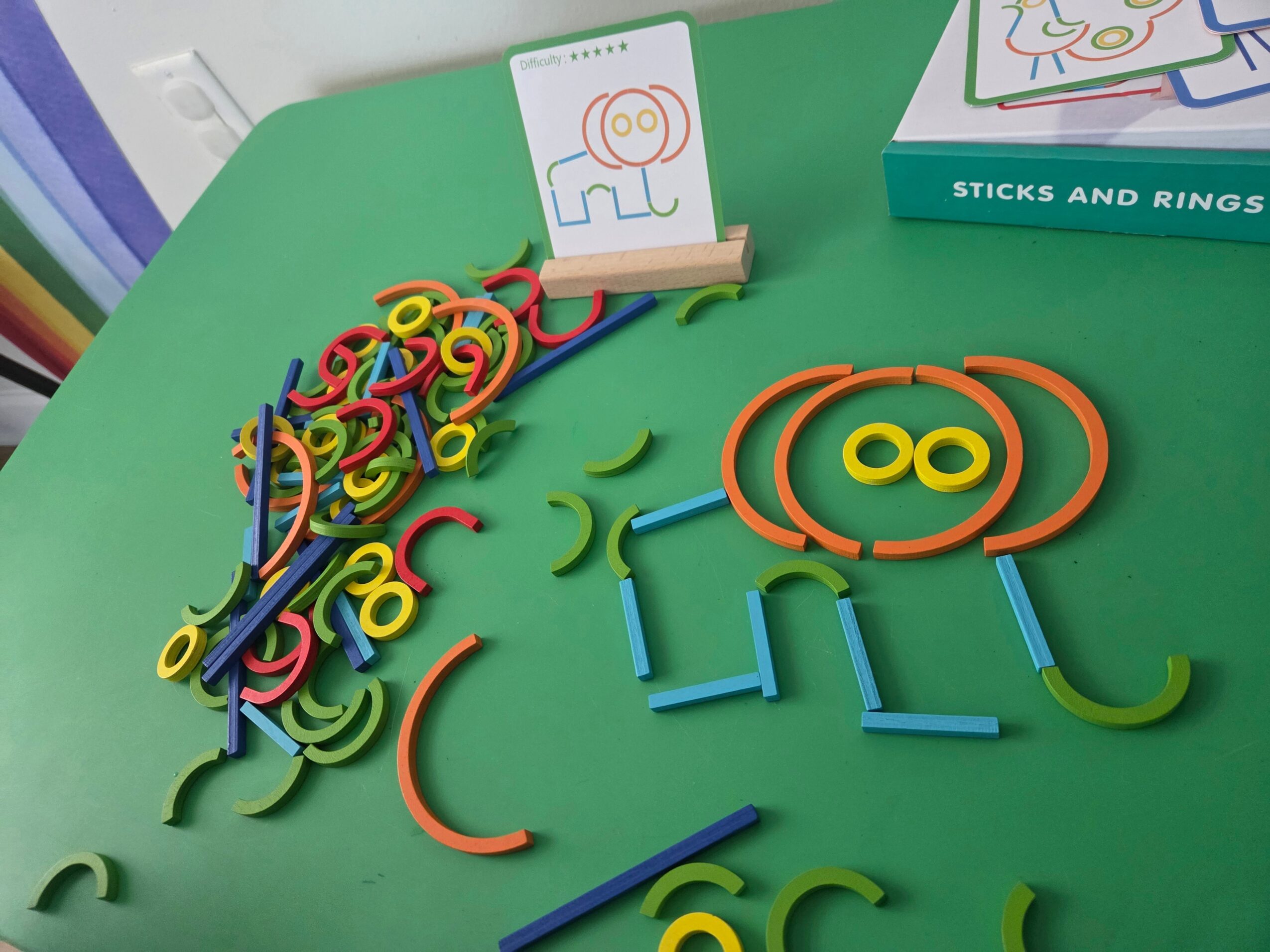 Colorful plastic sticks and rings scattered on a green surface with a partially visible box and a small stand with a drawing.