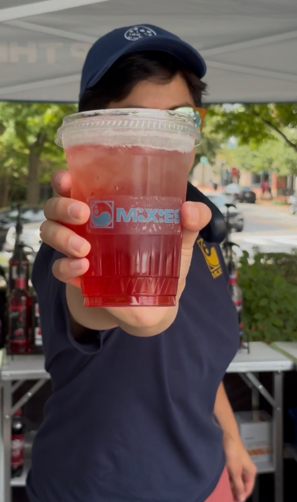 Person holding a large clear cup with red beverage, outdoors with trees and tents in background.