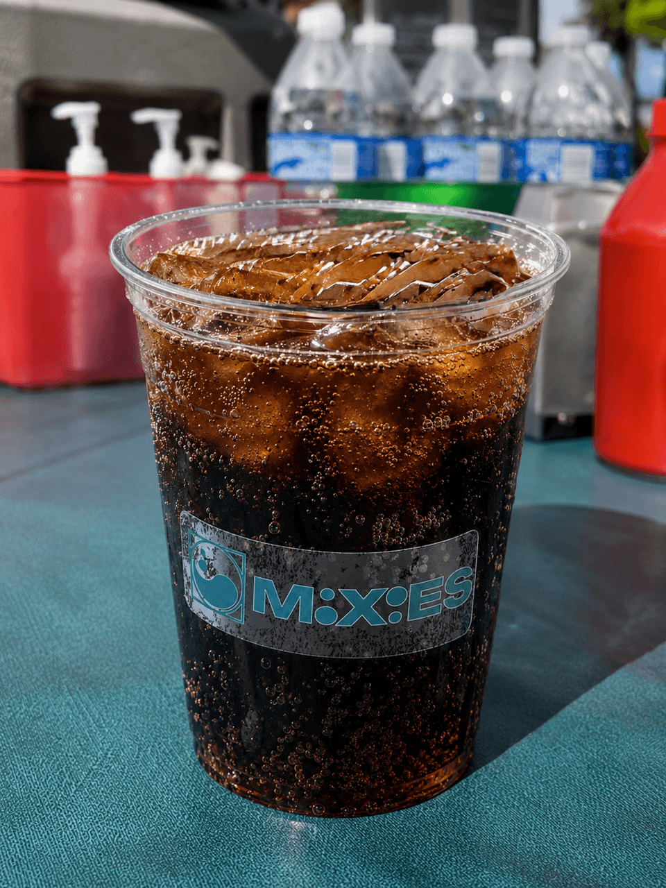 A plastic cup filled with dark soda and ice cubes, placed on a table with water bottles in the background.