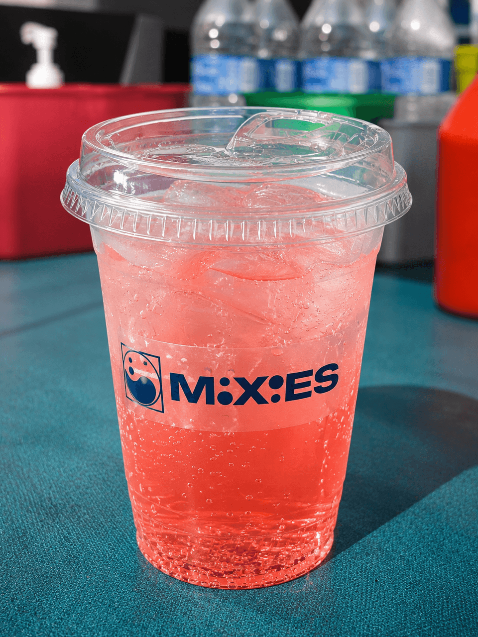 Clear plastic cup with pink beverage and ice, on a teal surface, with bottles and containers in background.