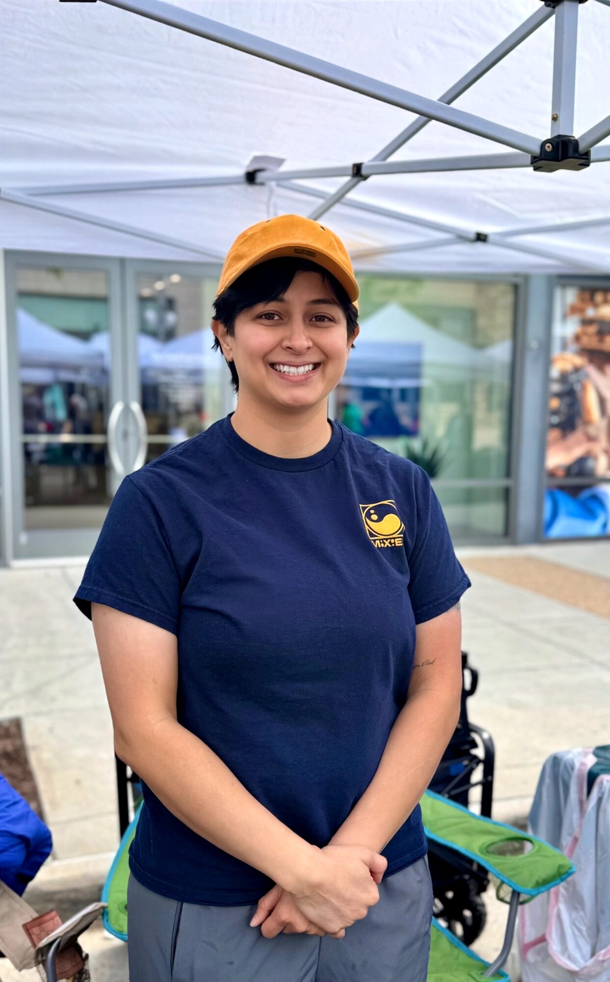 Smiling person wearing a navy T-shirt and orange cap standing outdoors under a canopy, with a building in the background.