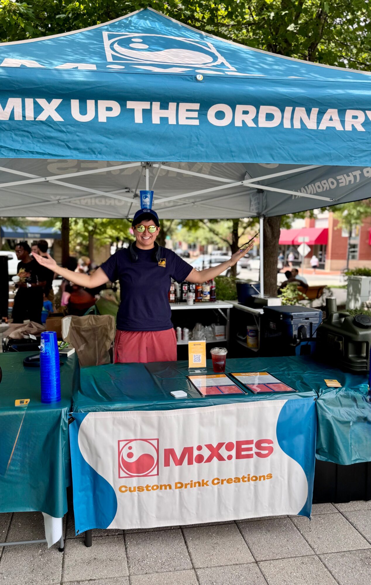 Person standing behind a table under a blue canopy with the text 'MIX UP THE ORDINARY,' outdoors with trees and buildings in background.
