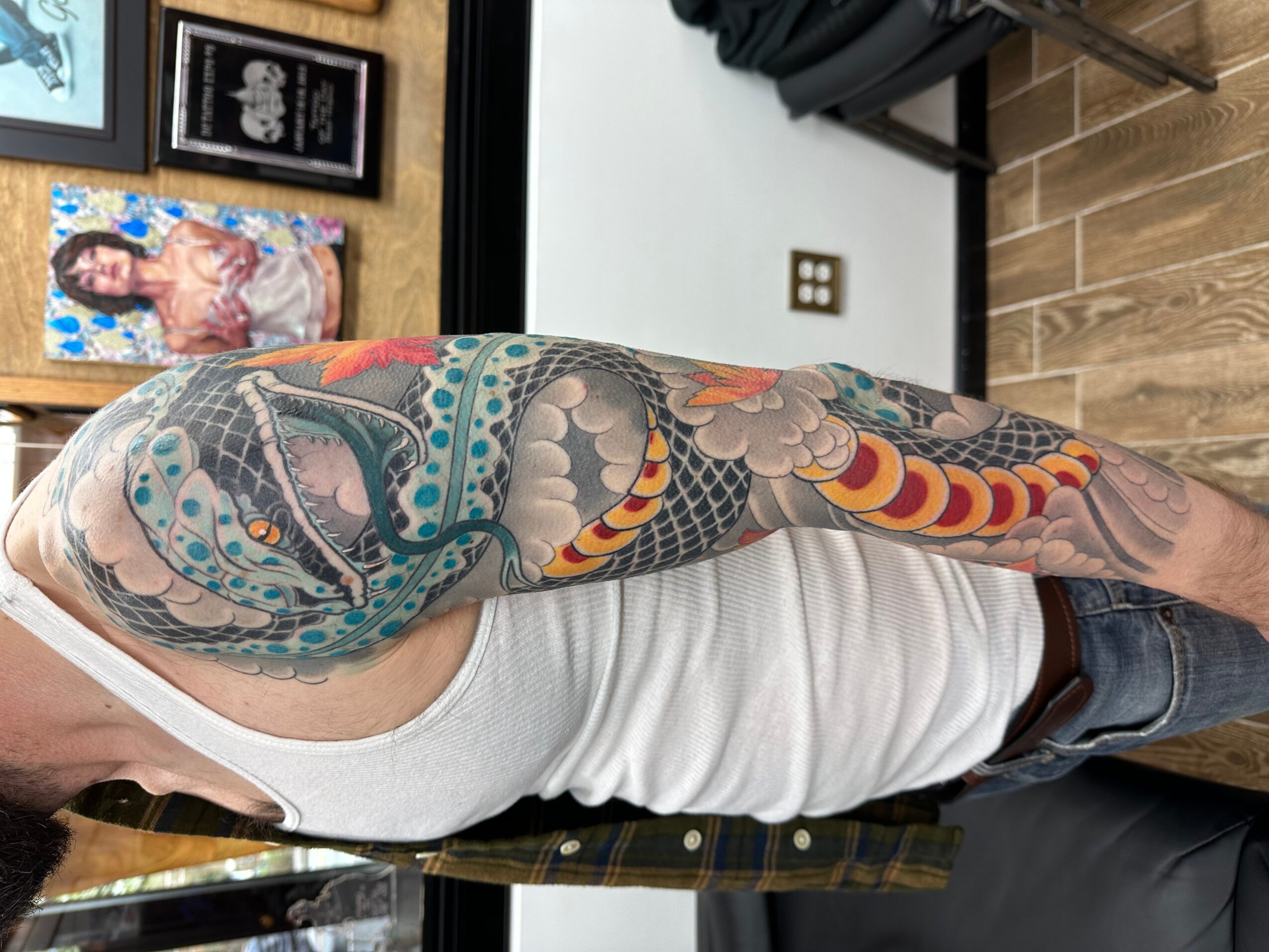 Person with colorful tattoo sleeve on arm, wearing white tank top, in indoor setting with framed pictures on wall