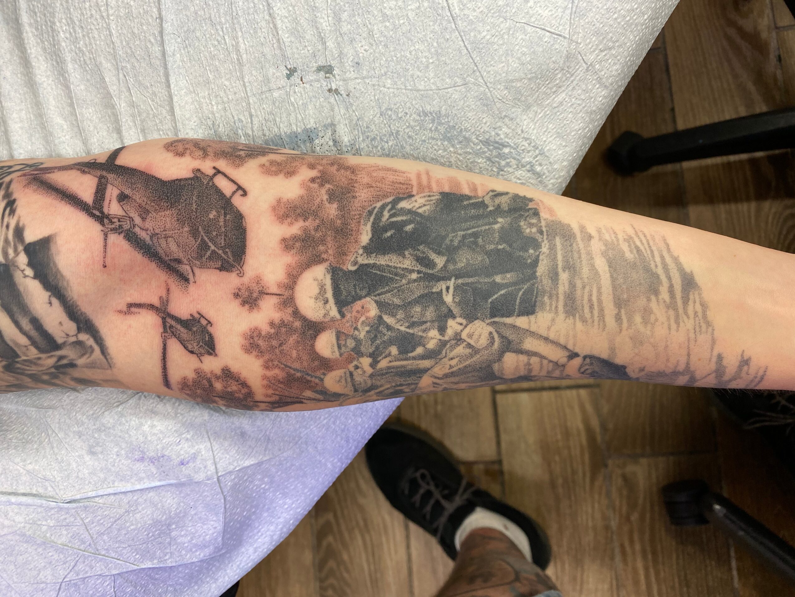 Tattooed arm with a detailed black and gray scene of people and trees, resting on a light-colored surface.