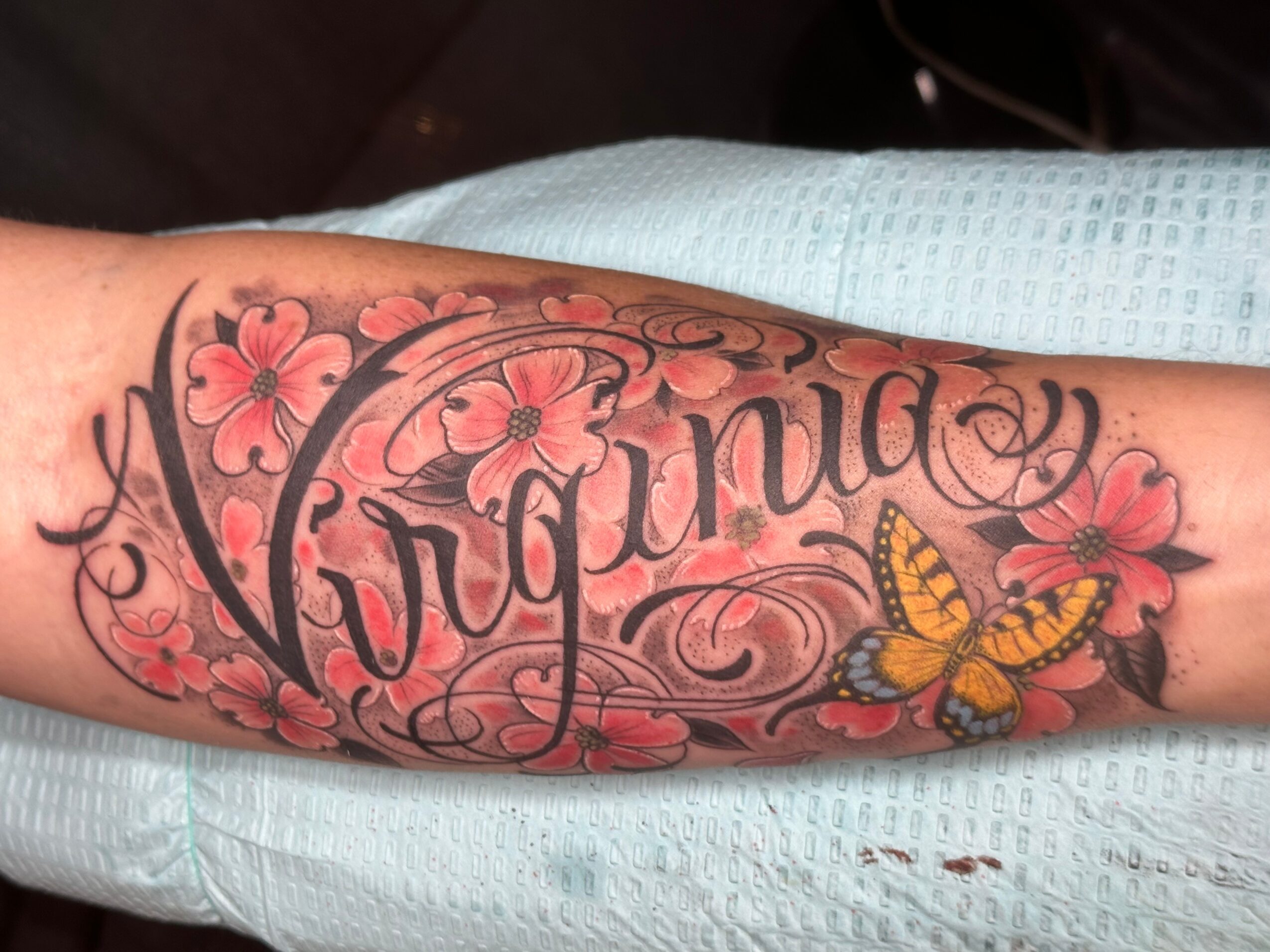 Tattoo of the word 'Virginia' with pink flowers and a yellow butterfly on a person's arm.