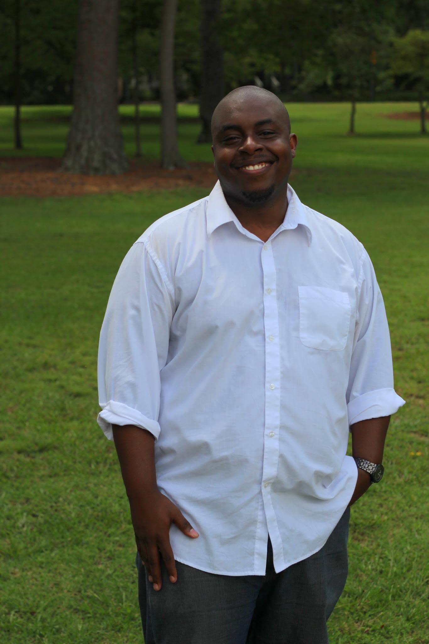 Smiling man in white shirt standing outdoors on grass with trees in background.