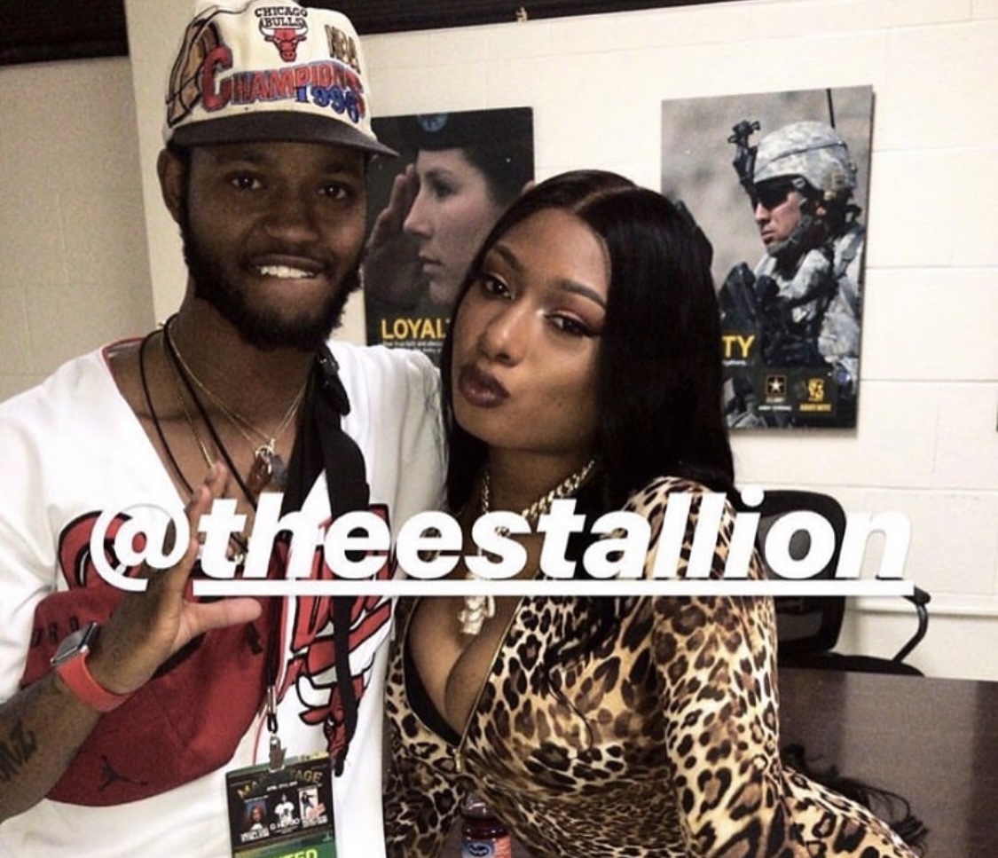 Two people posing for a photo, one wearing a cap and the other in a leopard print top, with posters in the background.