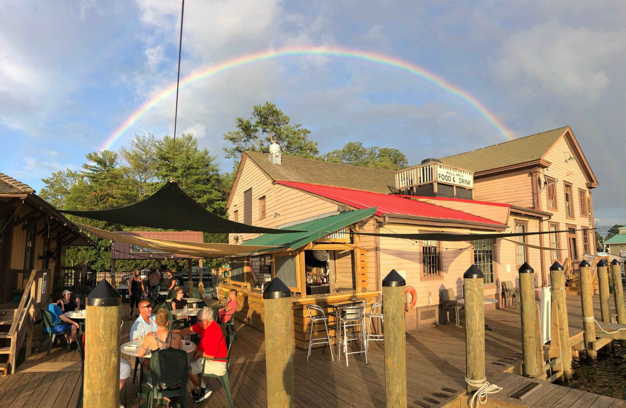 Rainbow over buildings and outdoor seating area with people, trees, and cloudy sky.
