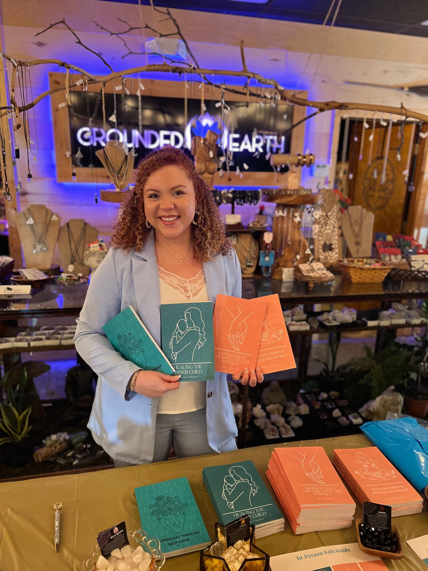 Smiling woman holding colorful books in a store with shelves and a sign in the background.