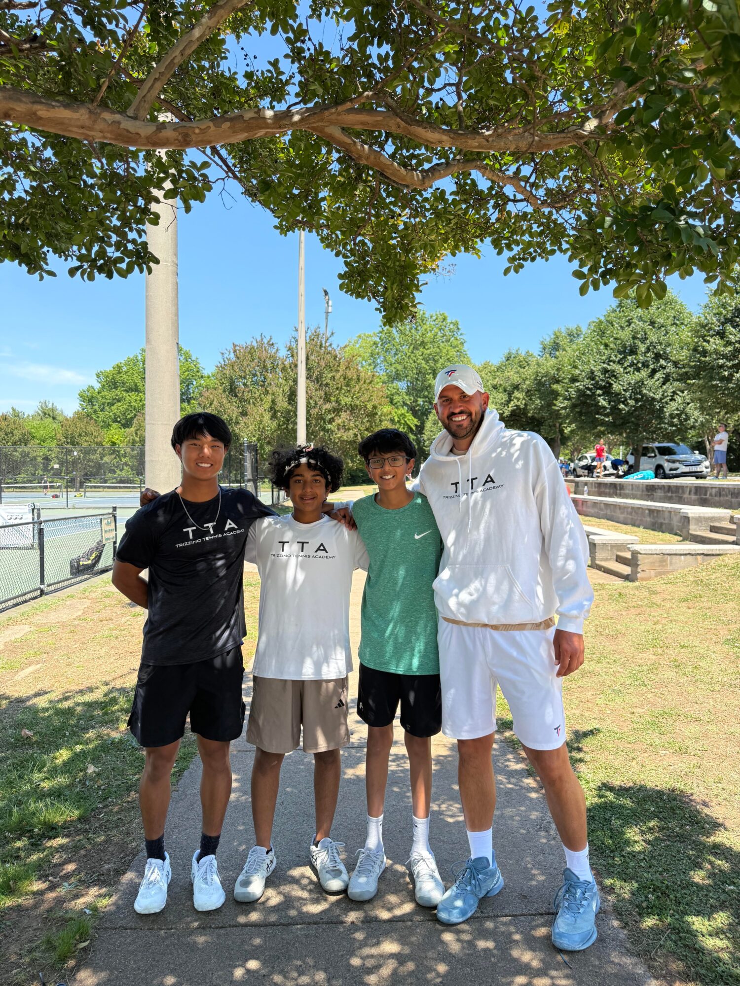 Four people standing outdoors under a tree, with a park and cars in the background.