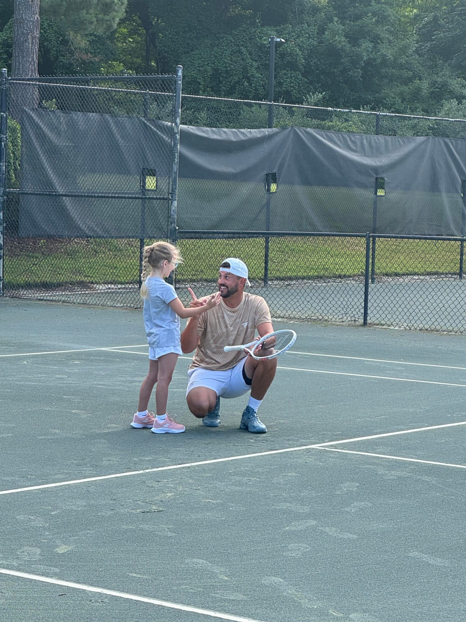 Adult kneeling on tennis court talking to young girl holding tennis racket, fence and trees in background.