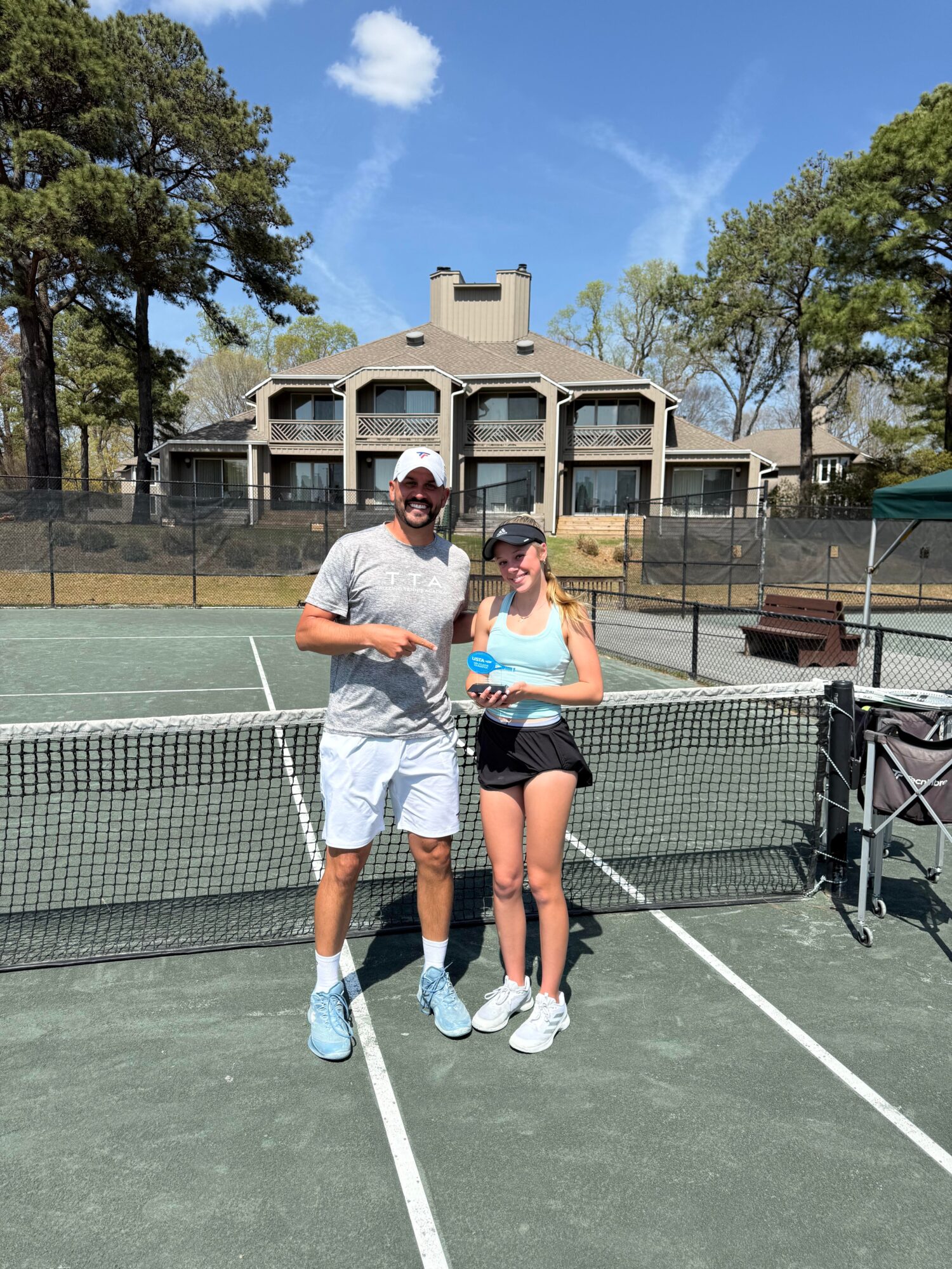 Two people standing on a tennis court holding a trophy, with a large building and trees in the background.