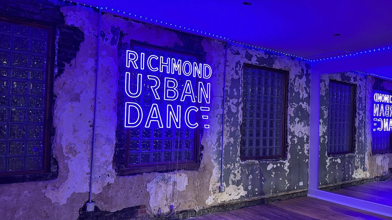 Neon sign reads 'Richmond Urban Dance' on a weathered wall with three windows, illuminated by blue lighting.