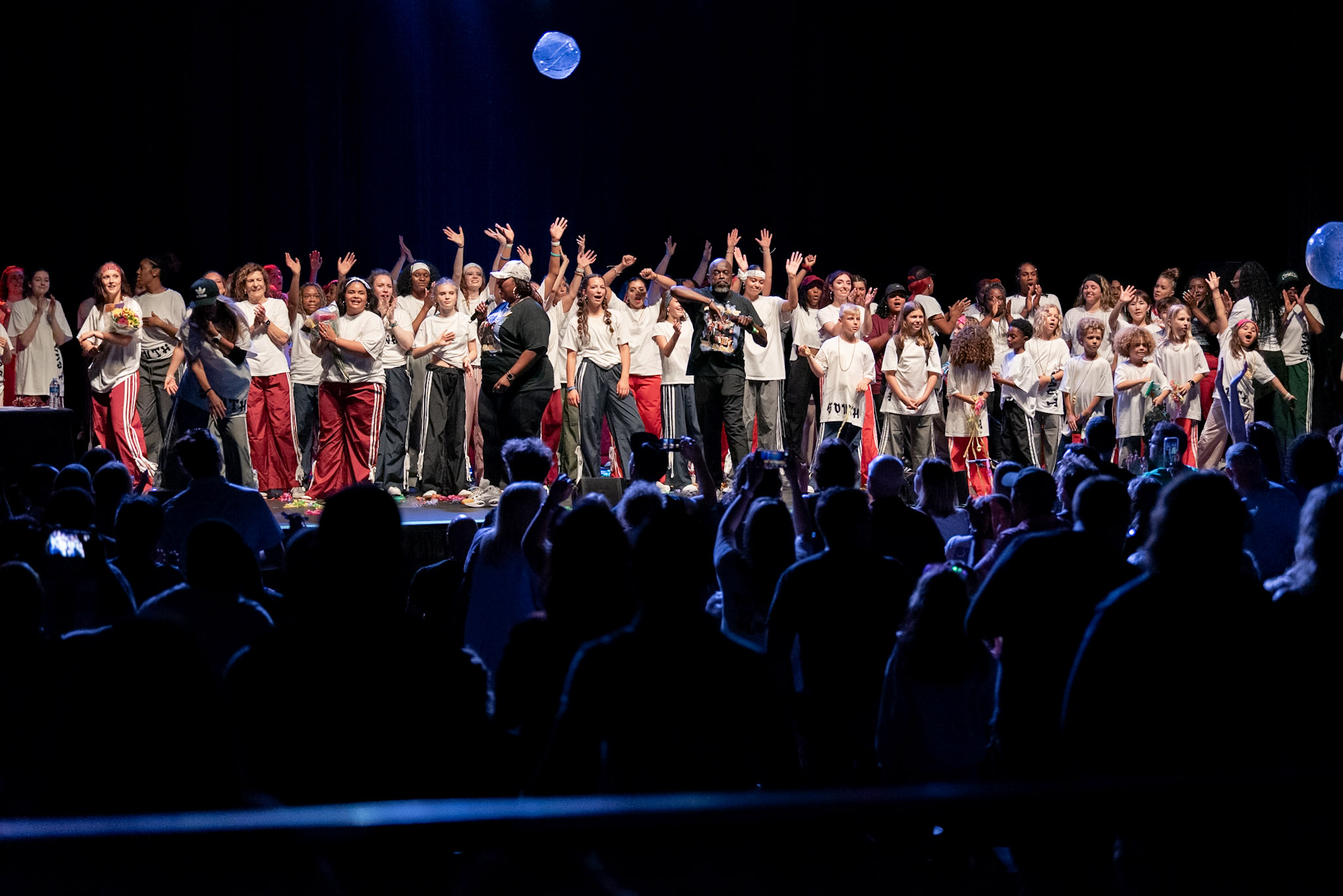Large group of children and adults on stage performing with balloons, audience watching in darkness.