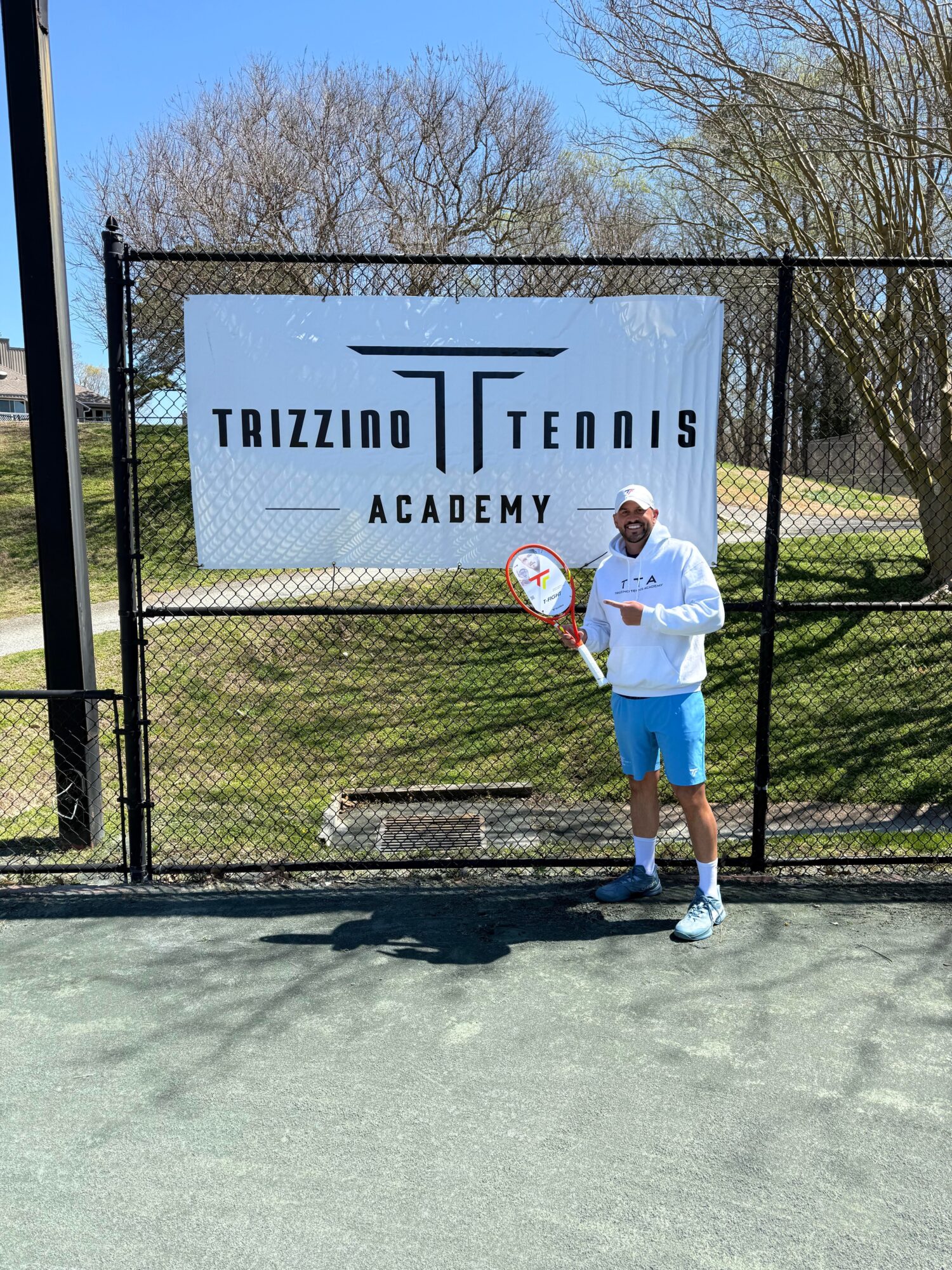 Person standing on tennis court holding a racket in front of a sign that reads 'Trizzino Tennis Academy' with trees in background.