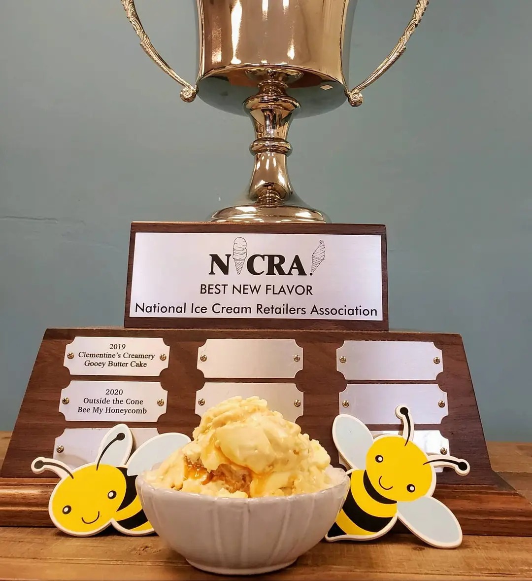 A bowl of ice cream in front of a trophy with a sign, surrounded by bee-shaped decorations, on a wooden surface.