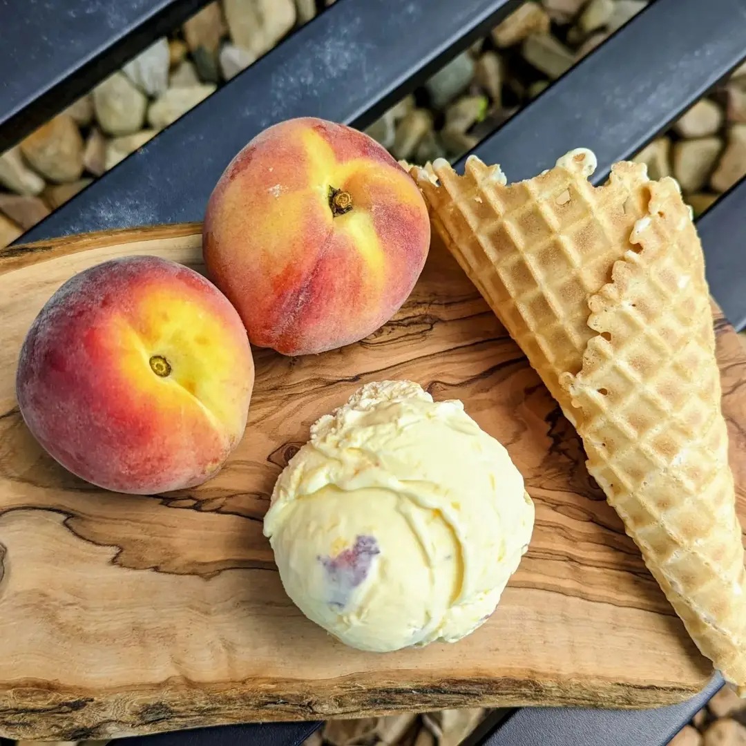 Two peaches, an ice cream scoop, and an ice cream cone on a wooden board outdoors.