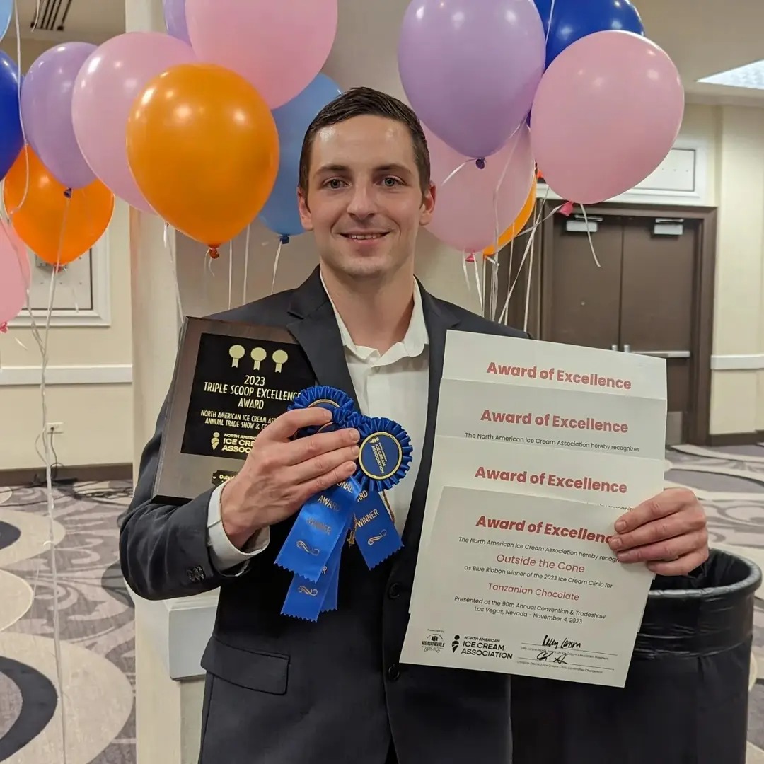 Man in suit holding award certificate and blue ribbon, smiling, balloons in background, indoor setting.