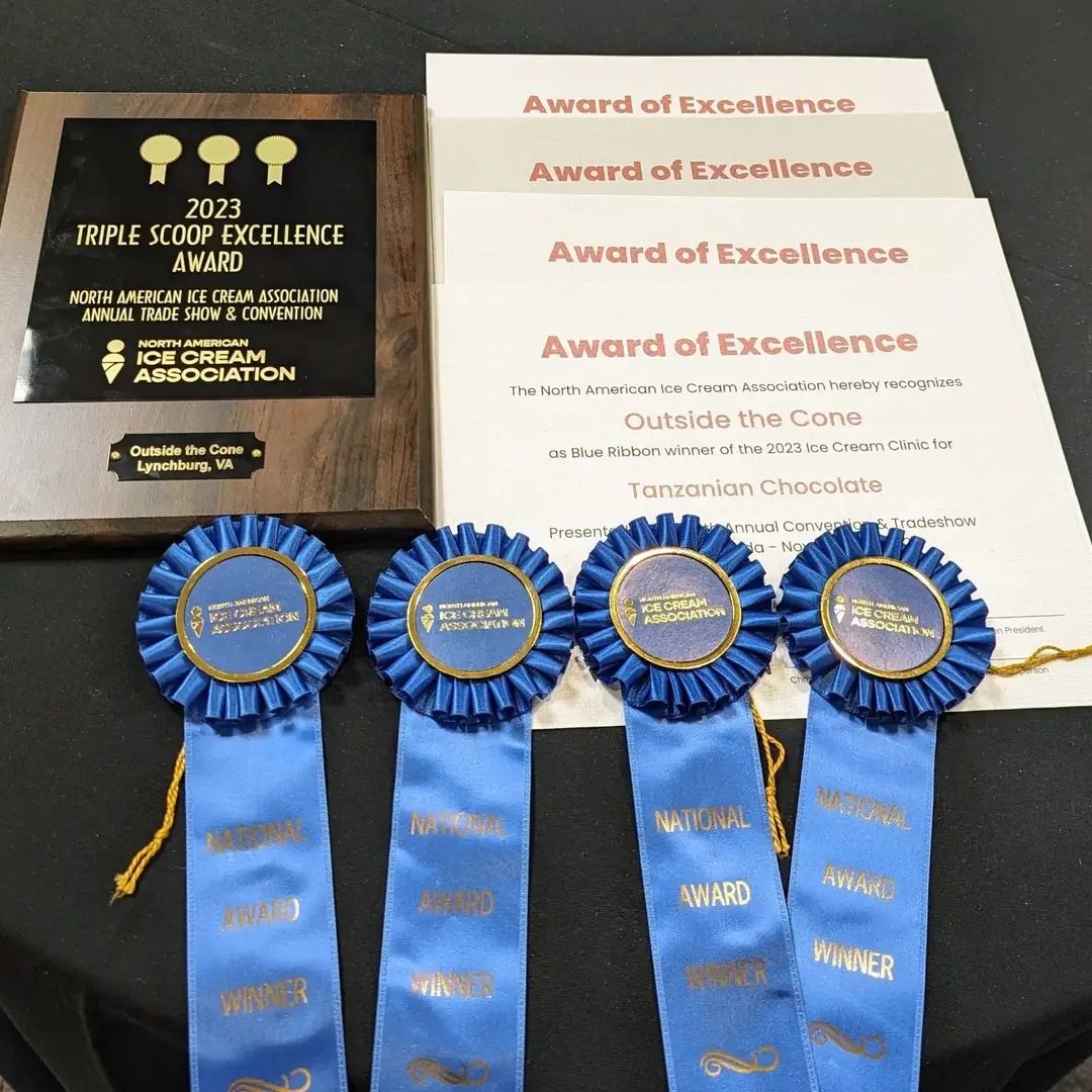 Four blue award ribbons with gold text and yellow strings, certificates, and a wooden plaque on a black surface.