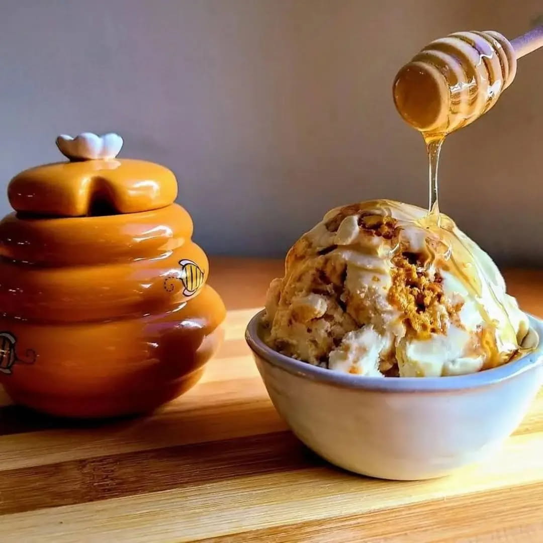 A bowl of ice cream with honey being drizzled on top, next to a decorative ceramic container on a wooden surface.