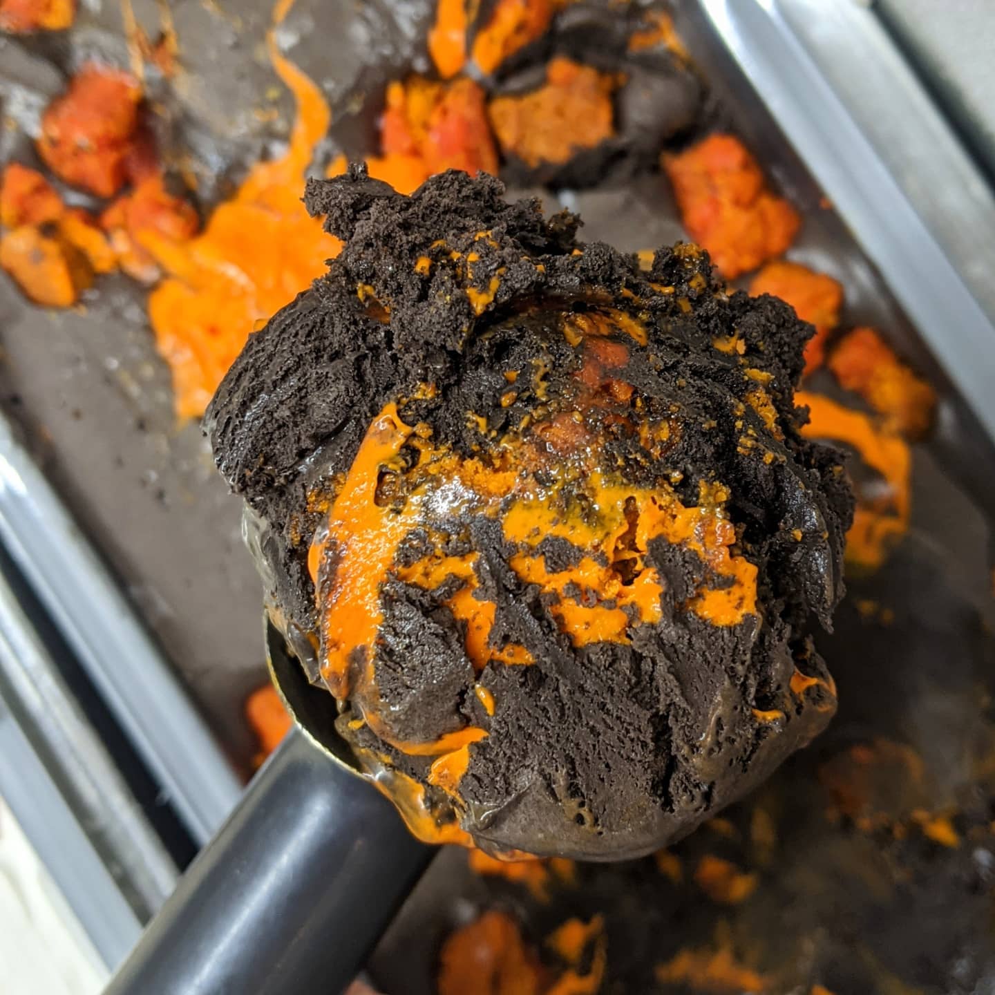Scooped dark, crumbly substance with orange patches, in a metal container, with more of the same in the background.