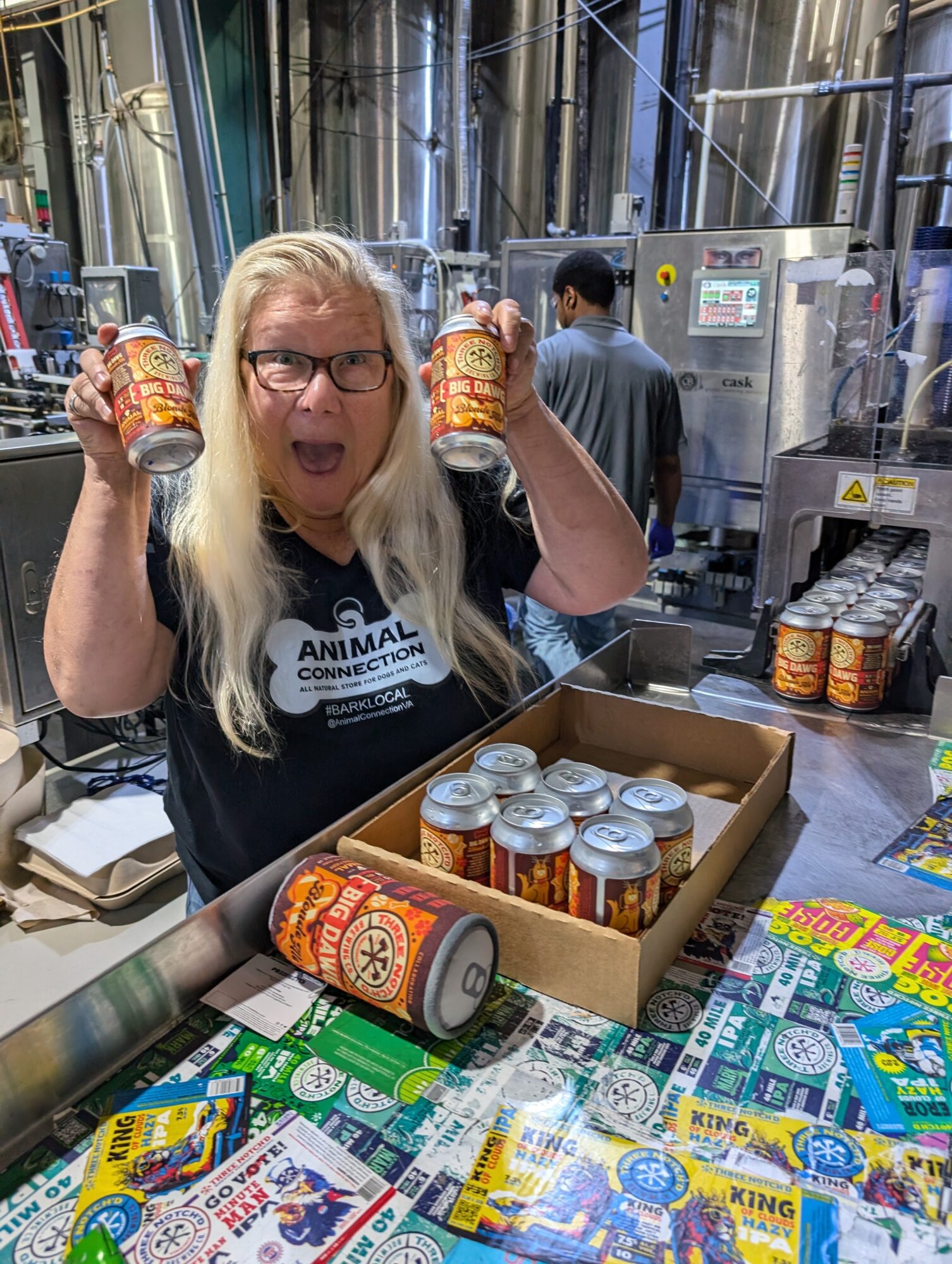 Woman with glasses holding two cans in a factory setting, surrounded by cans and colorful packaging on the table.